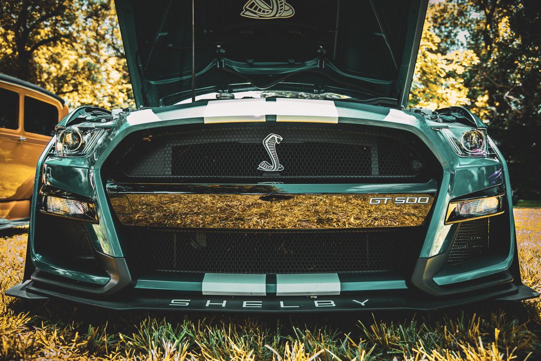 Front view of a teal Shelby GT500 Mustang with hood open, showcasing the iconic cobra emblem and racing stripes.