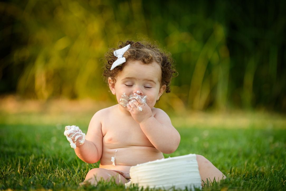 Baby girl with curly hair eating birthday cake smash on green grass outdoors.