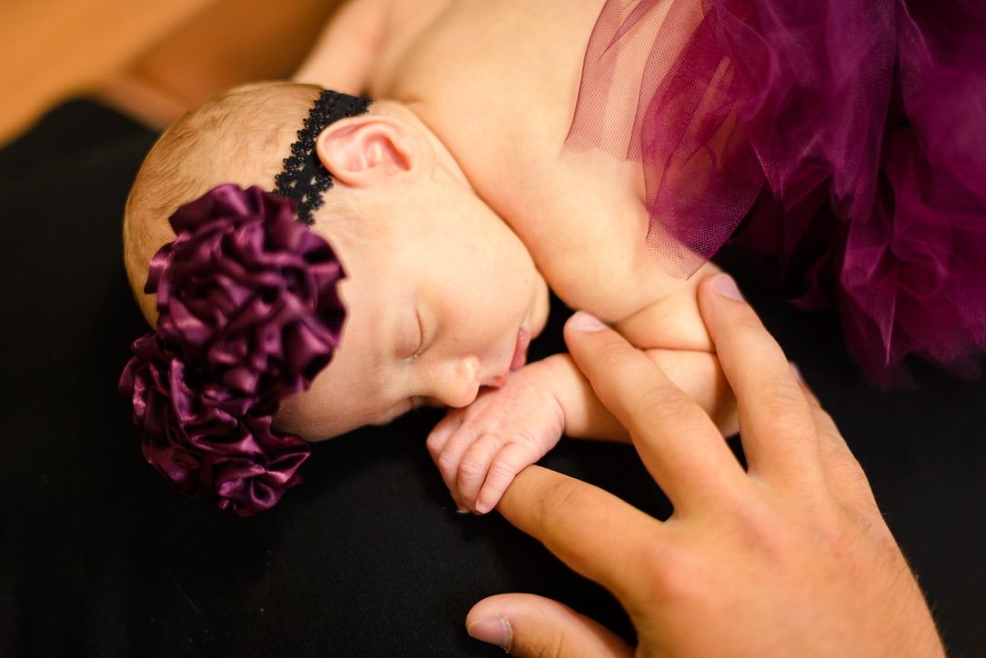 Sleeping newborn baby wearing a purple flower headband, cradled in adult hands against a black background.