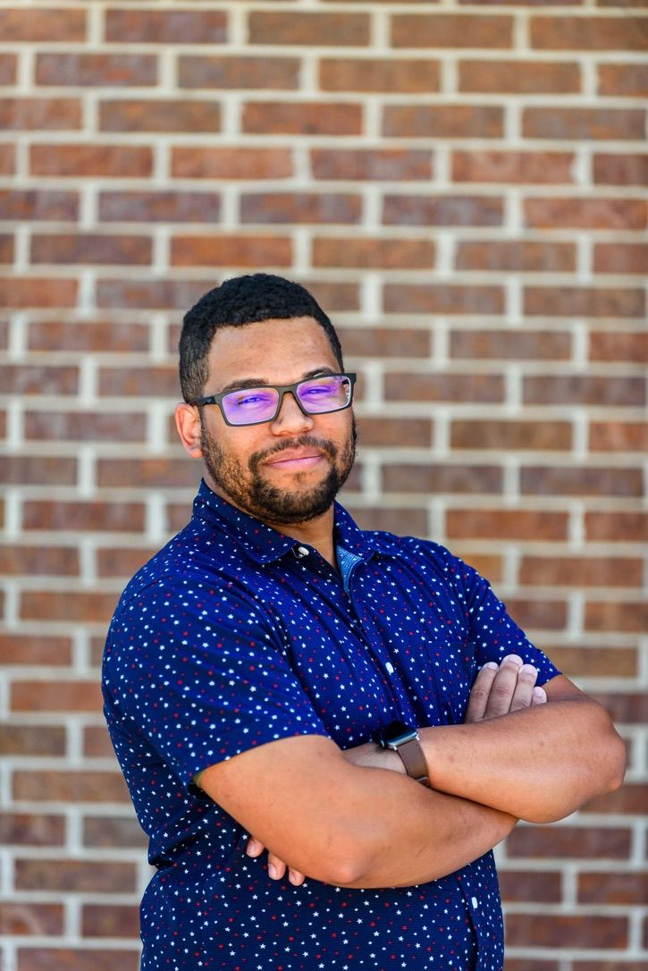 A confident bearded man with glasses poses arms crossed in a blue polka dot shirt against a brick wall.