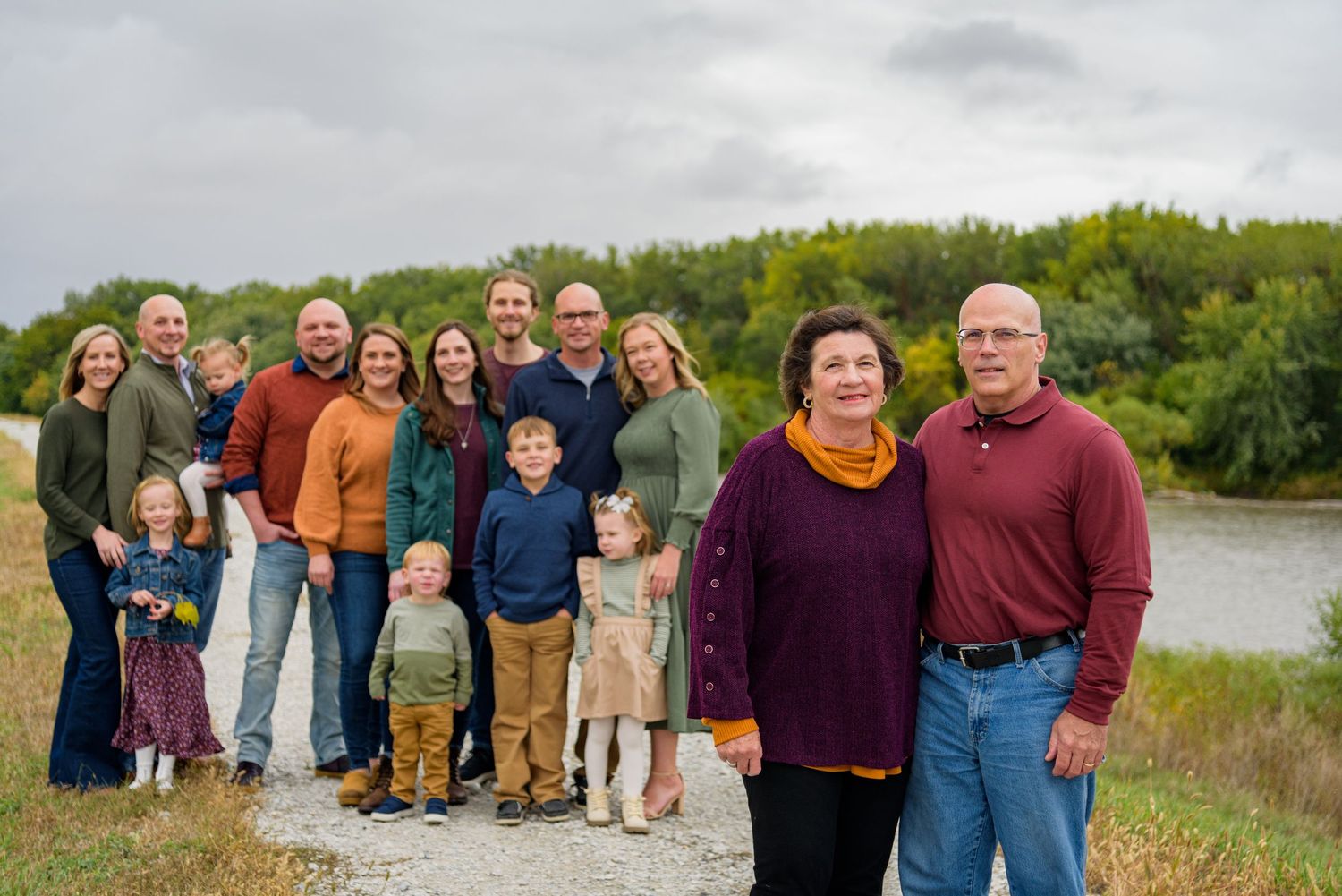Large family group portrait outdoors near a river with fall foliage, featuring multiple generations posing on a gravel path.