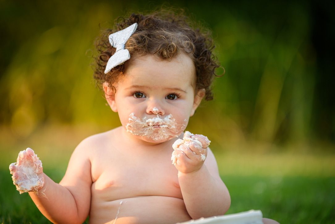 Adorable baby girl with curly hair eating cake with her hands outdoors during a first birthday smash cake session.