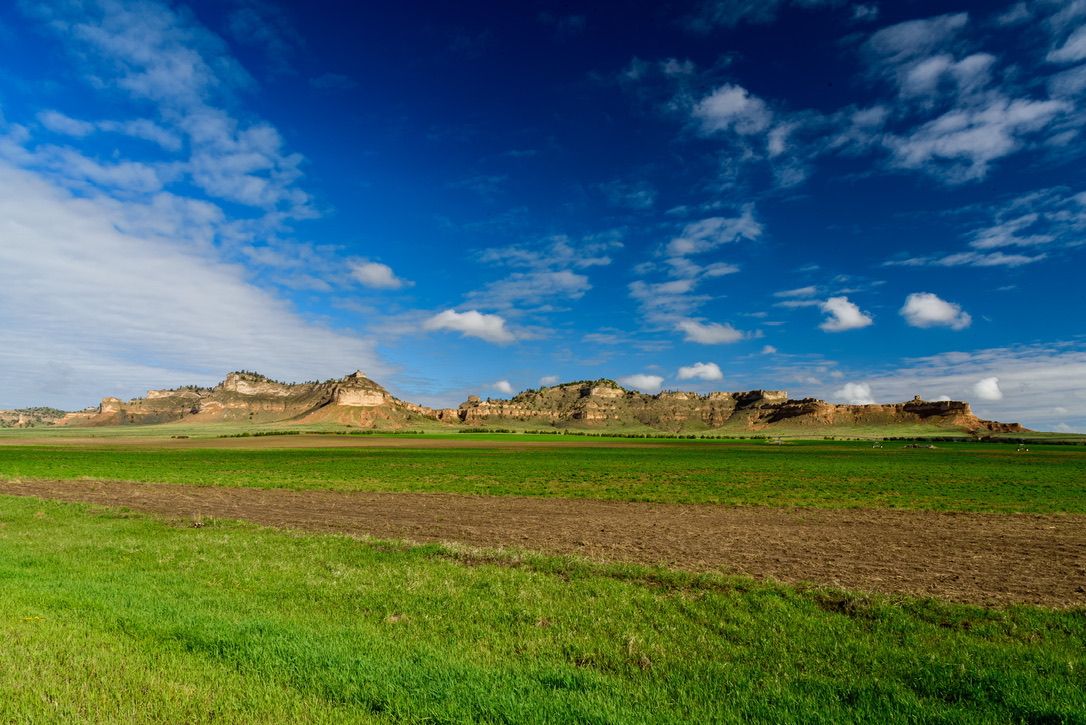 Green fields stretch before rugged sandstone buttes under a vivid blue sky with scattered clouds in a vast prairie landscape.