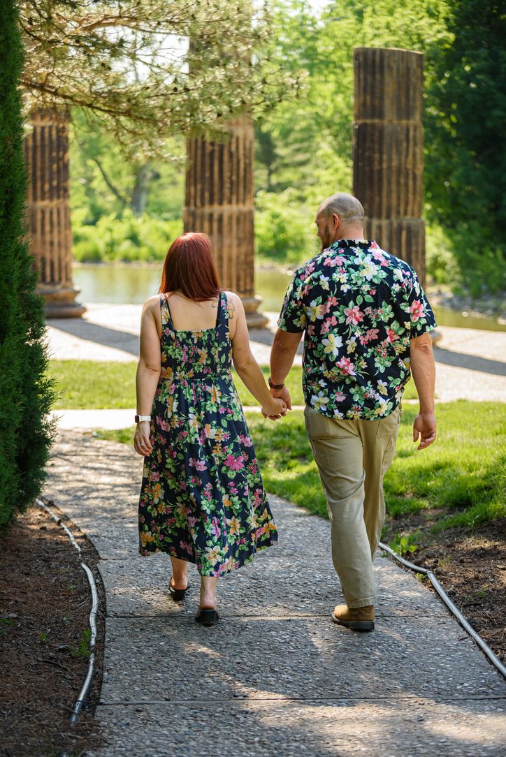Couple in matching floral outfits holding hands walking along a garden path with stone columns and lush greenery.