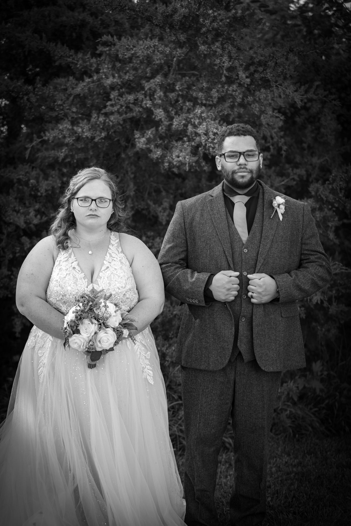 Black and white wedding photo of bride holding bouquet and groom in tweed suit standing outdoors.