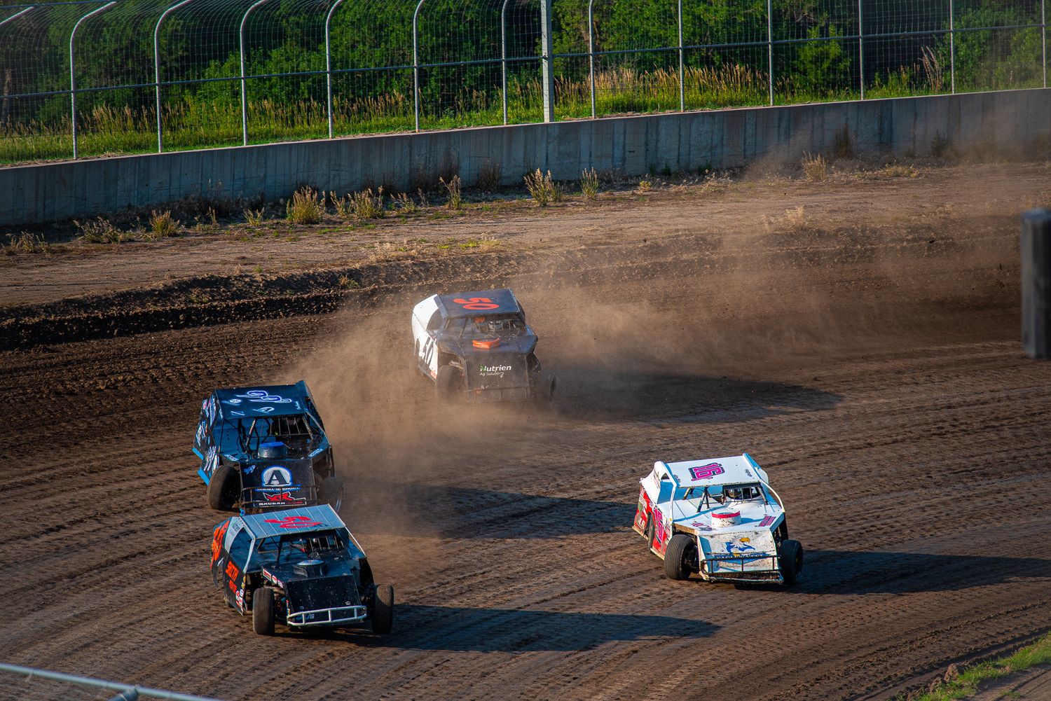 Four dirt track racing cars compete on a dusty oval circuit, kicking up clouds of dirt during an intense race.