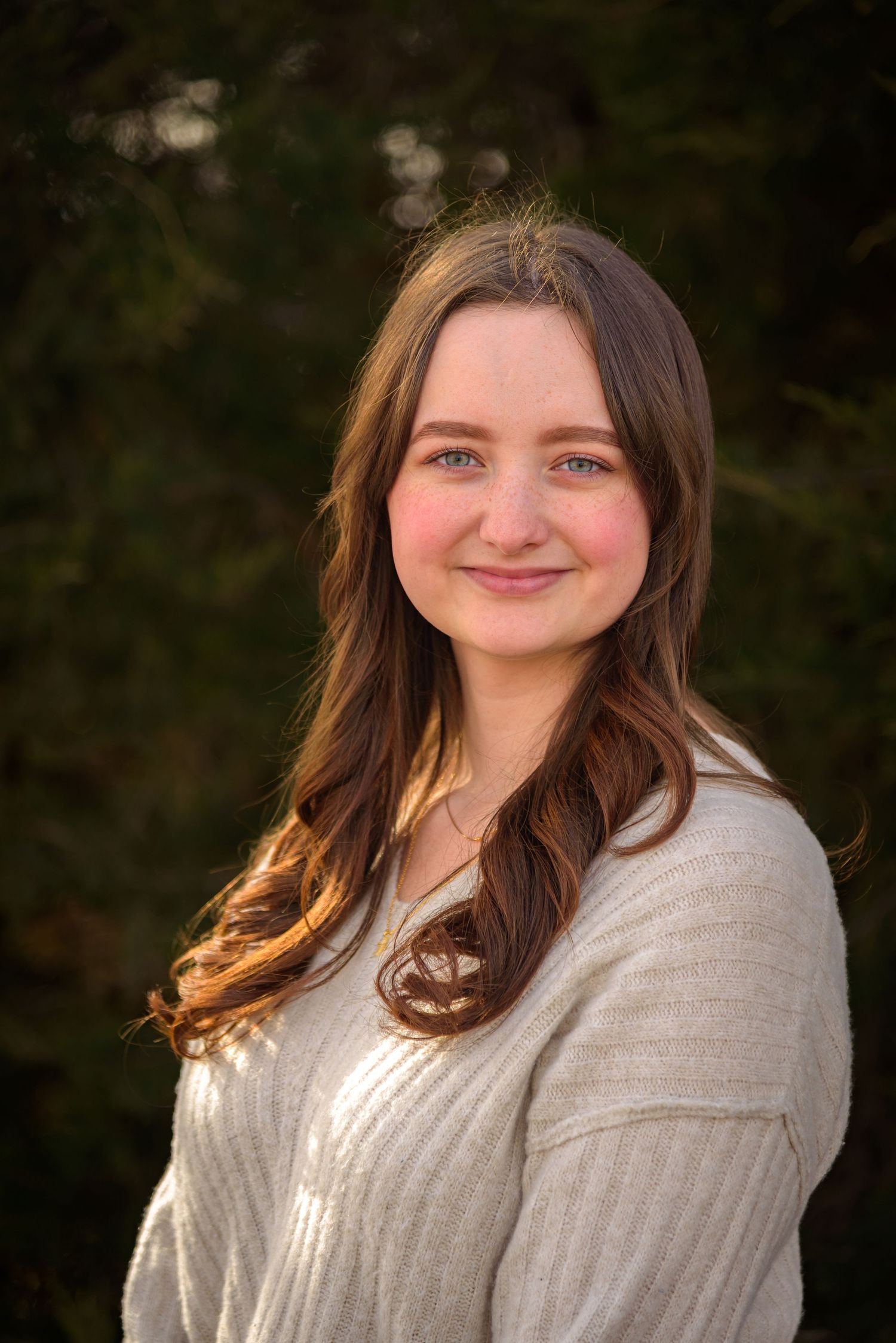 Young woman with long brown hair smiling outdoors in cream sweater with evergreen background. York, NE