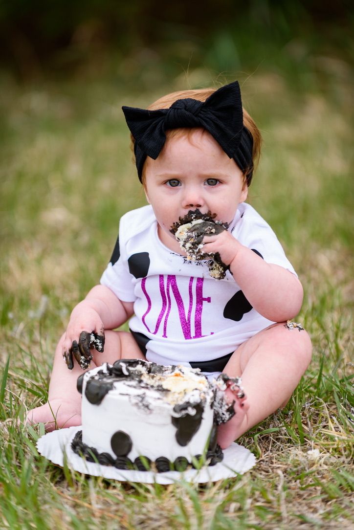 Baby girl in a black bow and ONE onesie smashing her first birthday cake outdoors on green grass.