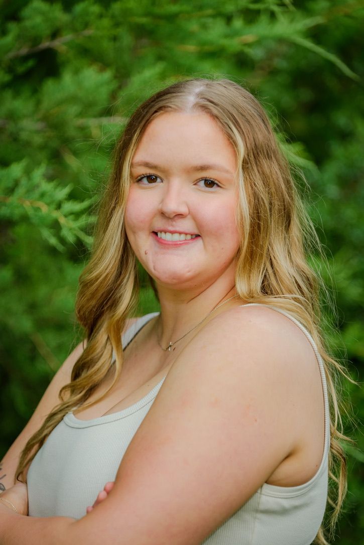 Smiling young woman with blonde wavy hair wearing a light tank top, posing outdoors with green trees in background.