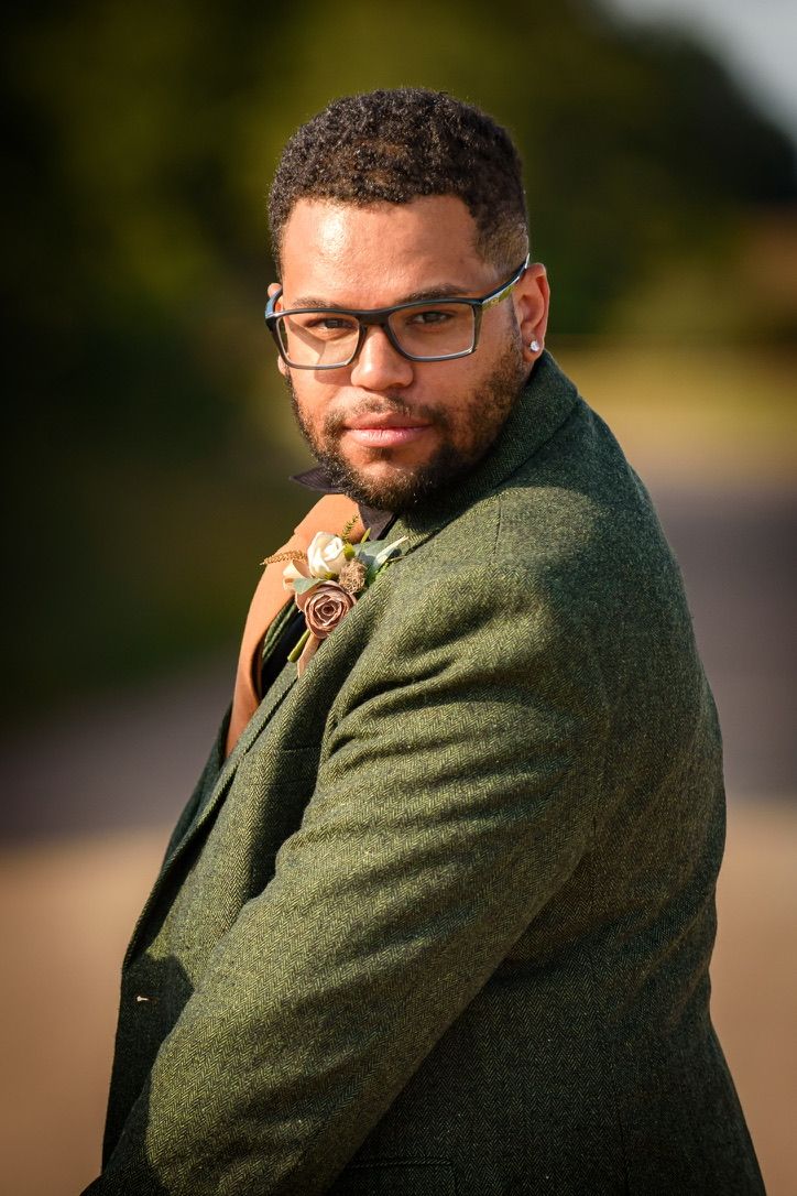 Groom in green tweed jacket with floral boutonniere poses outdoors, wearing glasses and a confident smile.