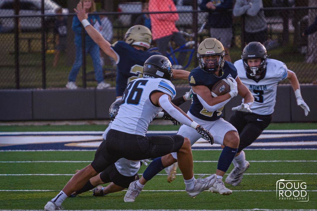 High school football player carries ball while two defenders in light blue jerseys attempt a tackle during a night game.