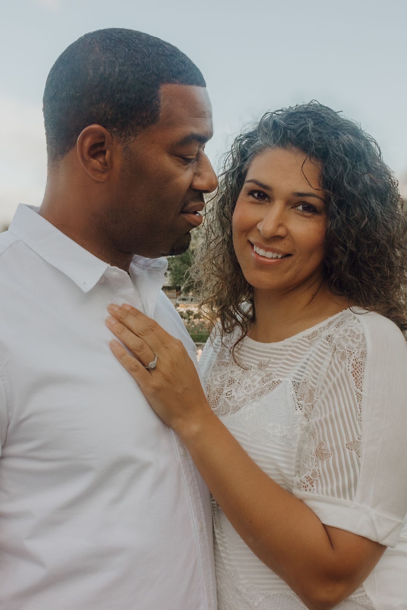 A smiling couple in white outfits share a tender moment outdoors, woman with curly hair rests hand on mans chest.
