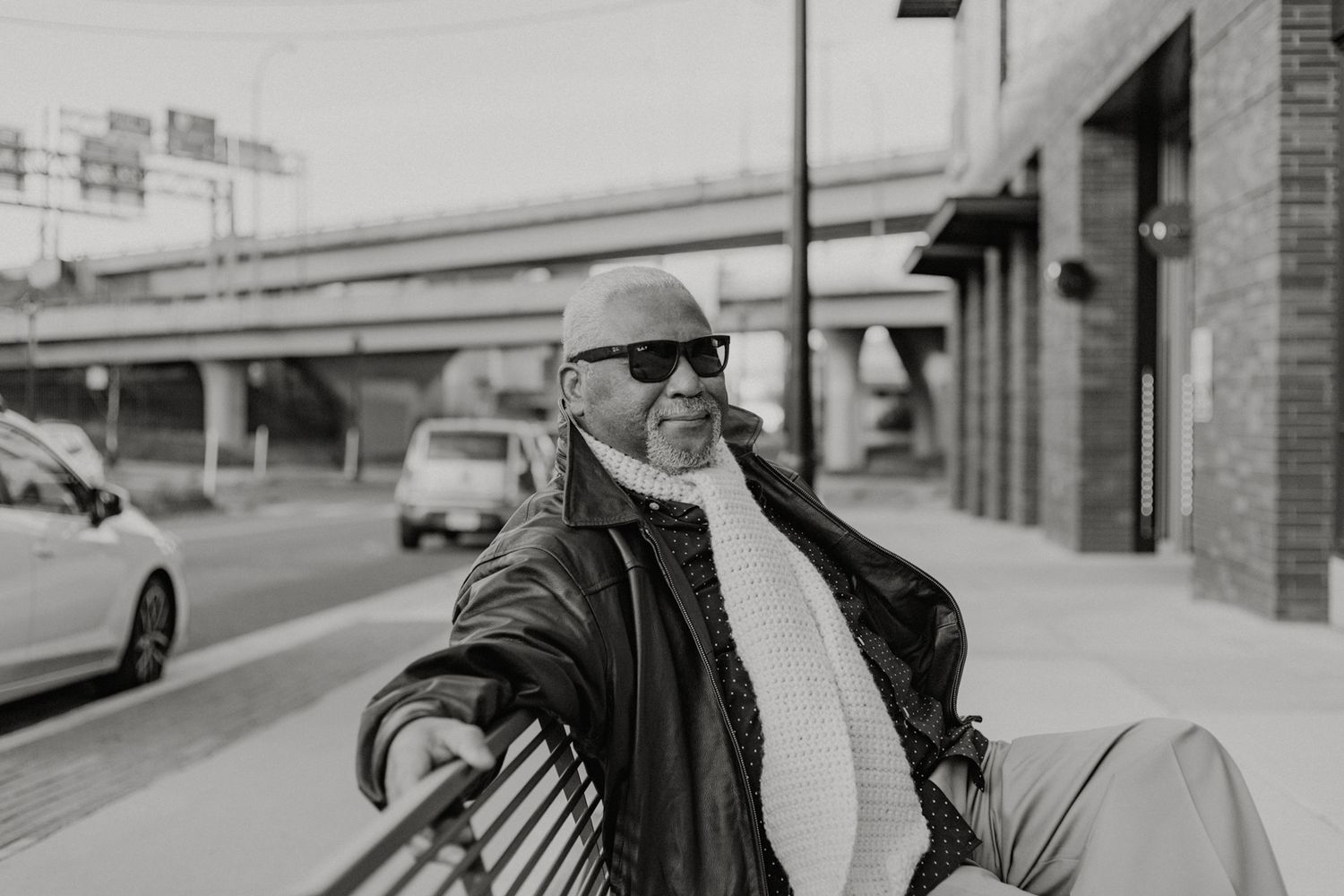 Elderly Black man in sunglasses and leather jacket relaxing on a bench near a city street overpass.