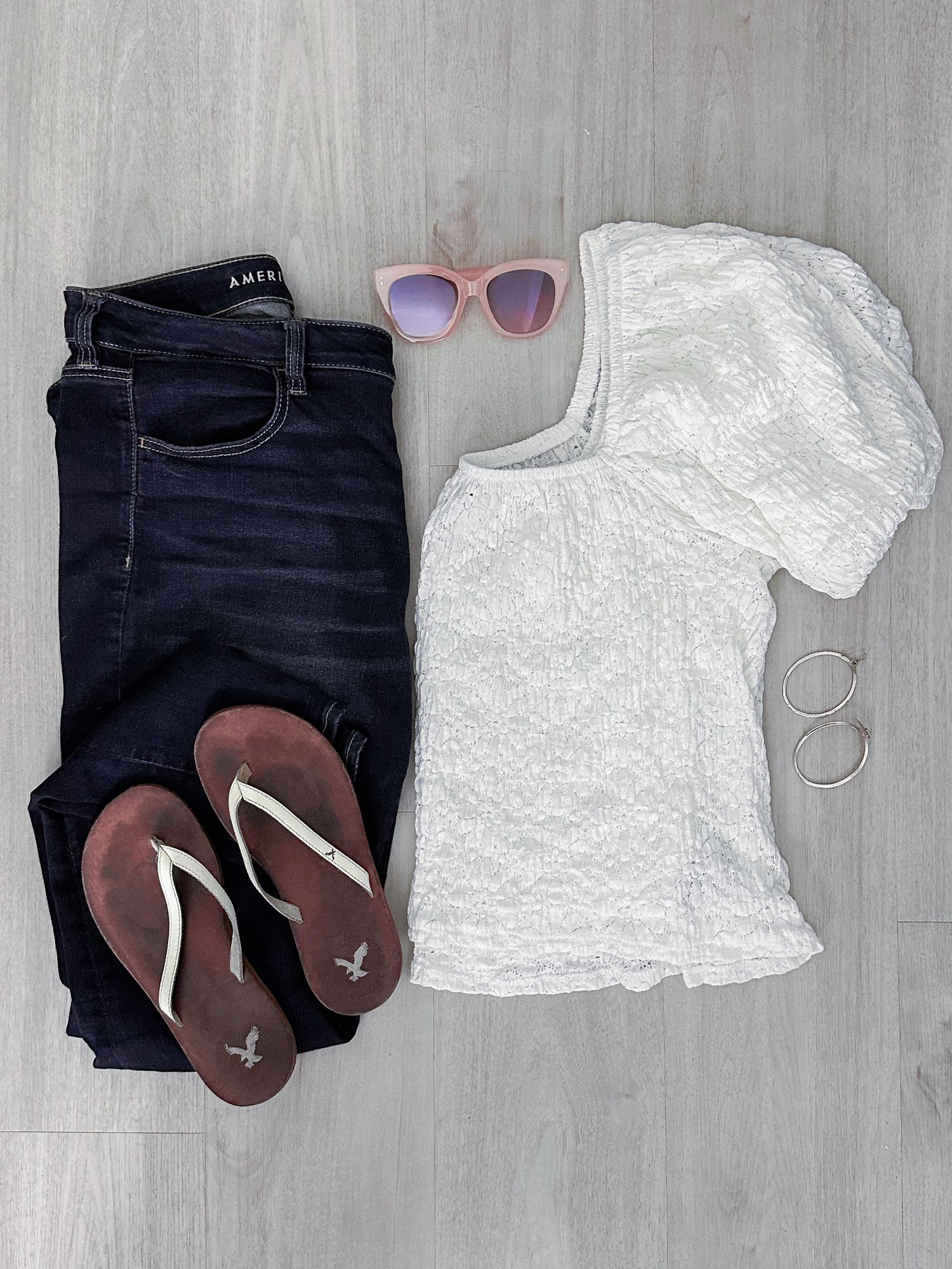 Flat lay outfit with dark jeans, white textured top, brown flip flops, pink sunglasses, and a silver ring.