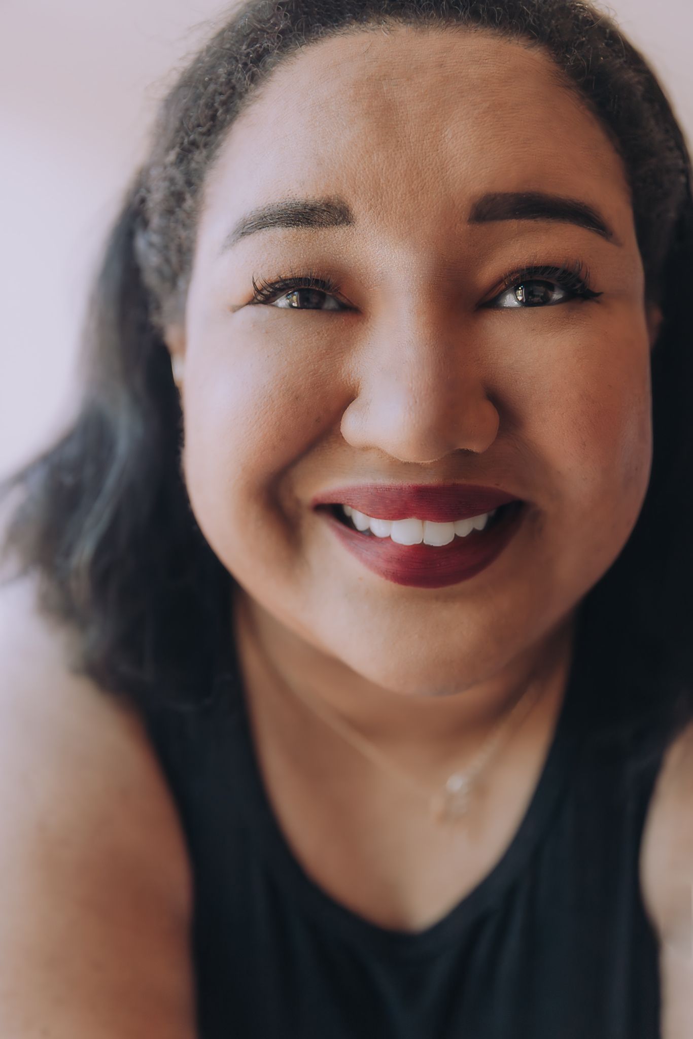 Smiling woman with dark hair and bold red lipstick wearing a black top and delicate necklace, close-up portrait.
