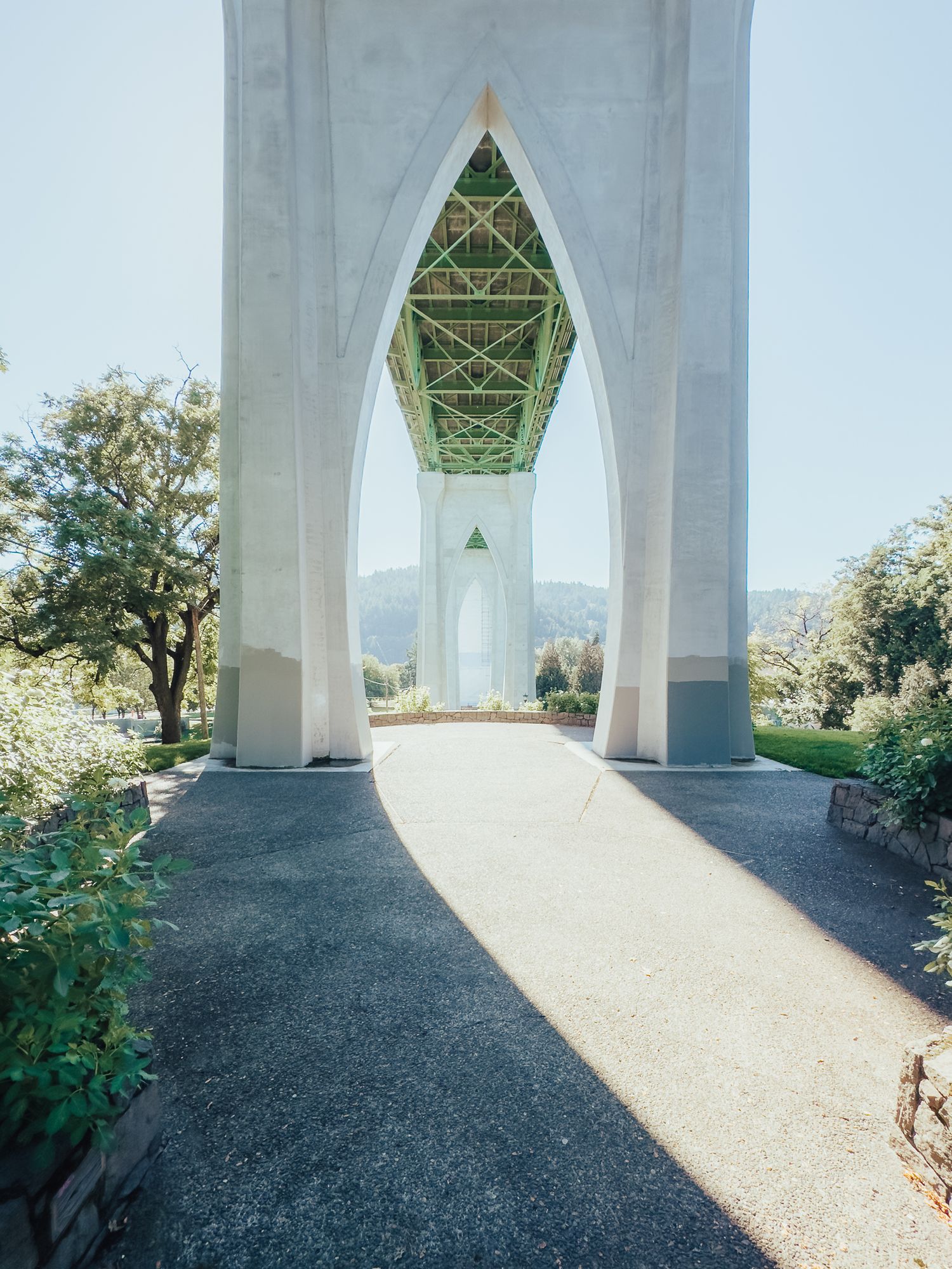 A paved path leads under the white Gothic arches of St. Johns Bridge in Portland, Oregon, surrounded by lush greenery.