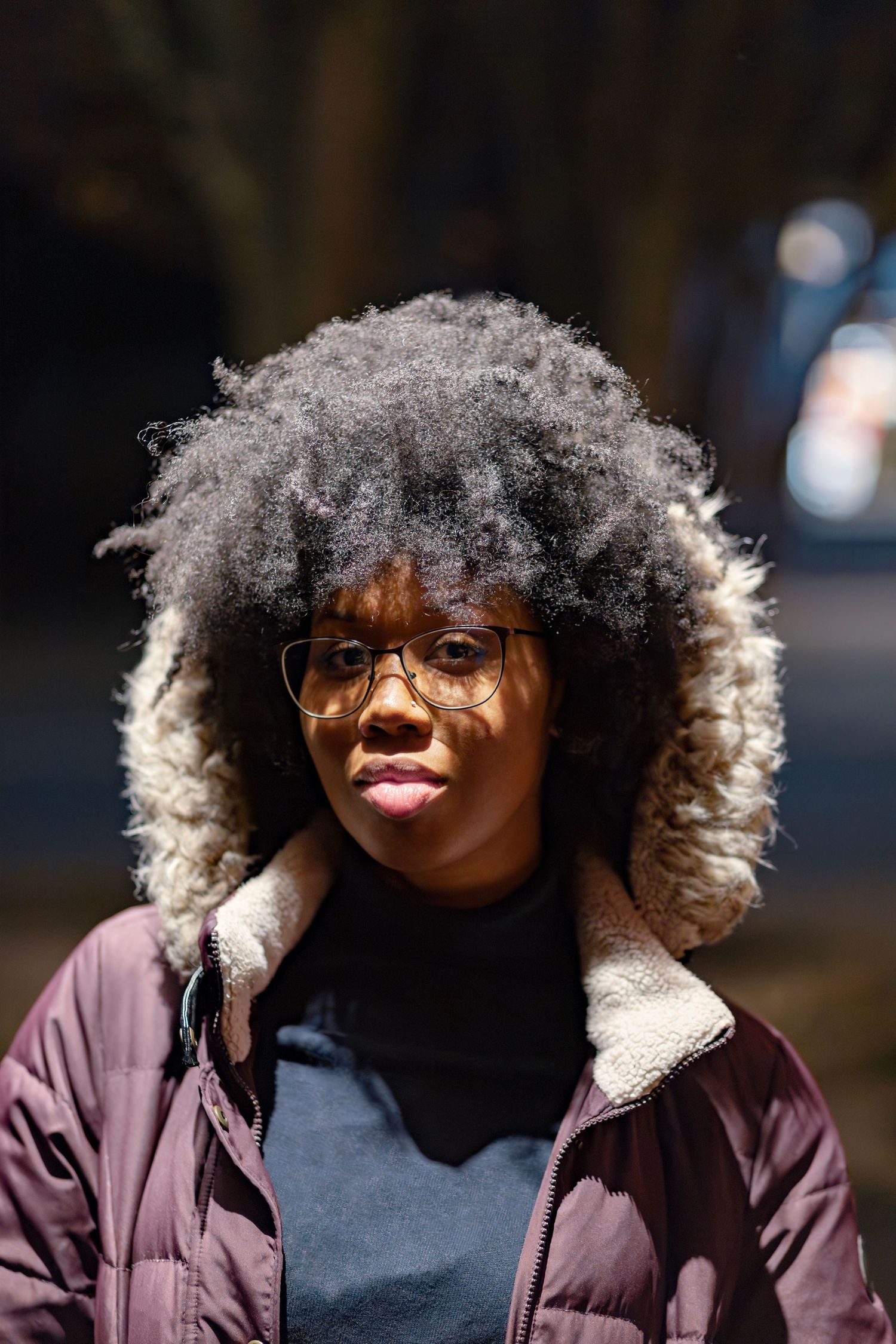 Young Black woman with natural afro hair and glasses wearing a purple winter jacket outdoors in sunlight.