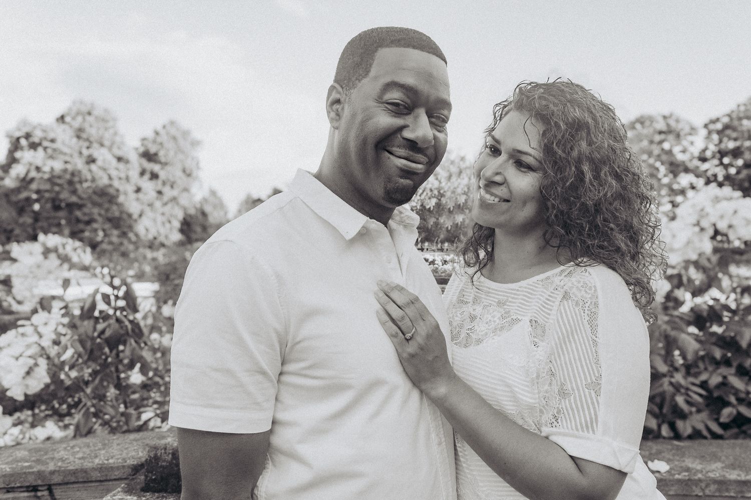 Black and white photo of a smiling couple embracing outdoors among blooming flowers in a garden setting.