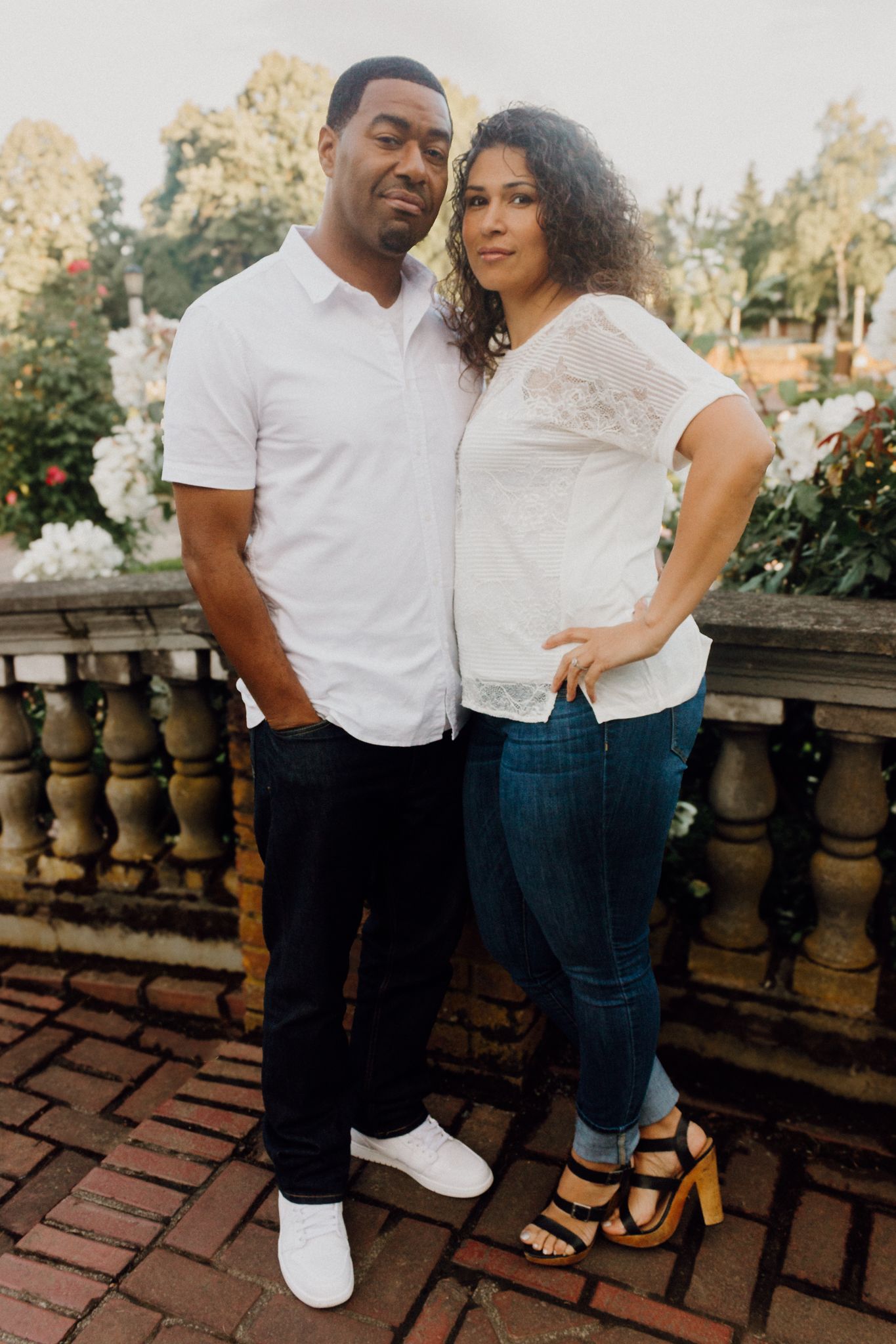 A couple in white tops and jeans poses together on a brick terrace surrounded by lush greenery and white flowers.