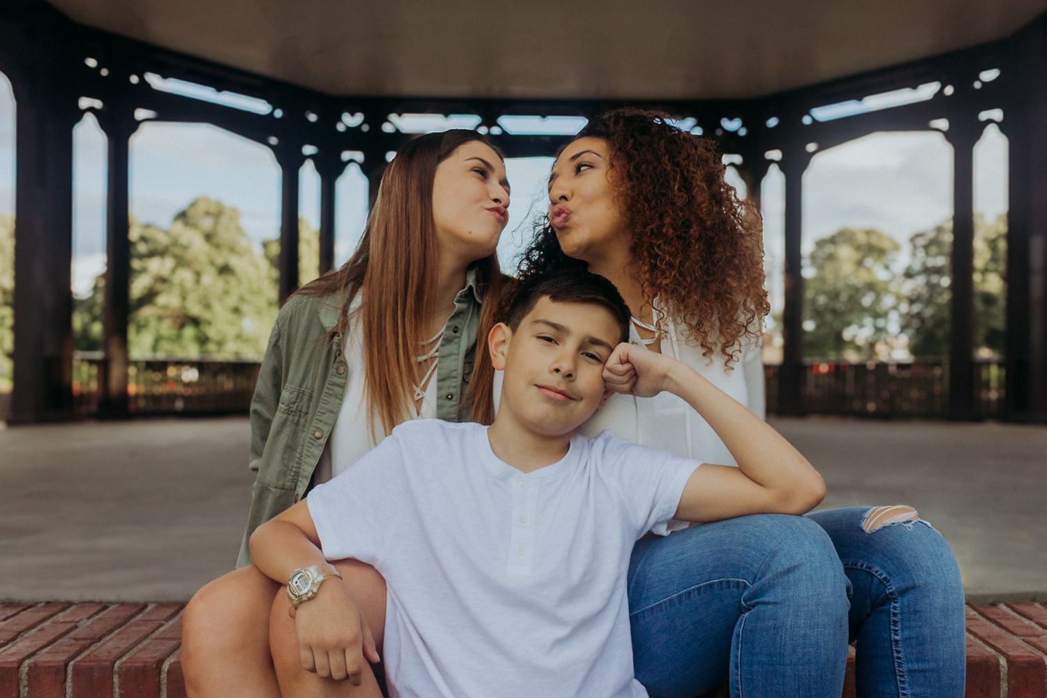 Two girls playfully kissing a boy's cheeks while sitting under a gazebo in a park.