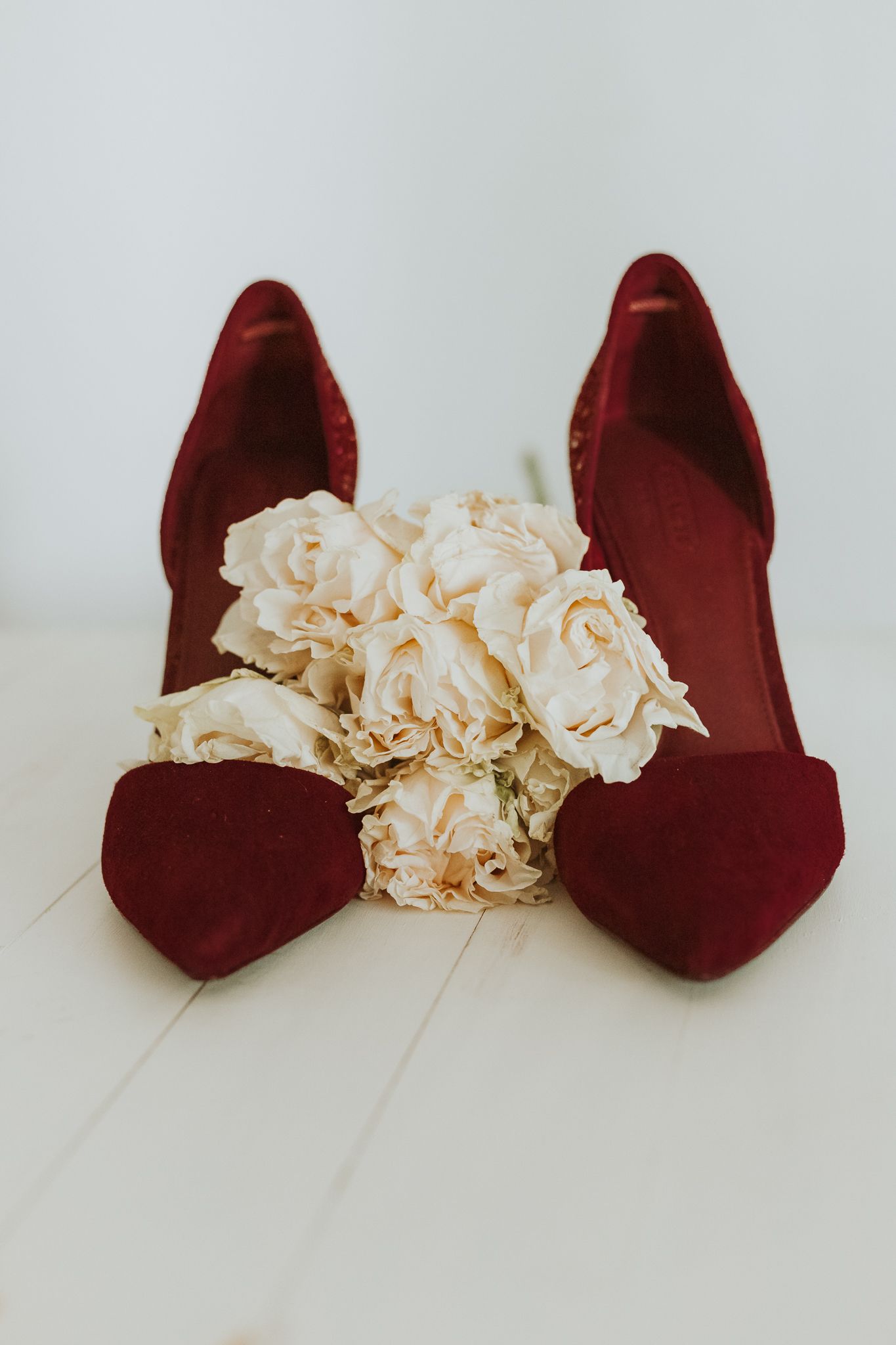 Elegant burgundy velvet heels displayed with cream roses bouquet on white surface for wedding bridal styling.