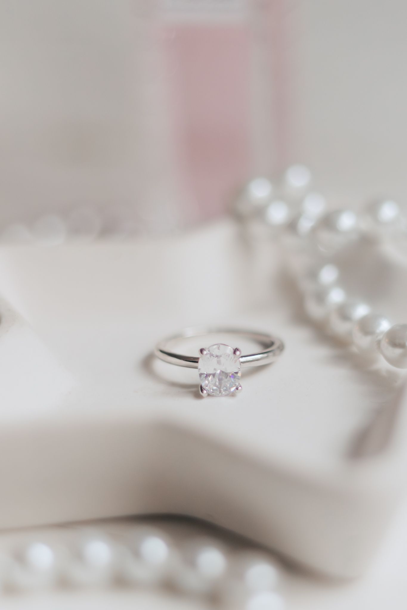 Elegant oval diamond solitaire ring in silver setting displayed on white dish with pearl necklace and pink perfume.