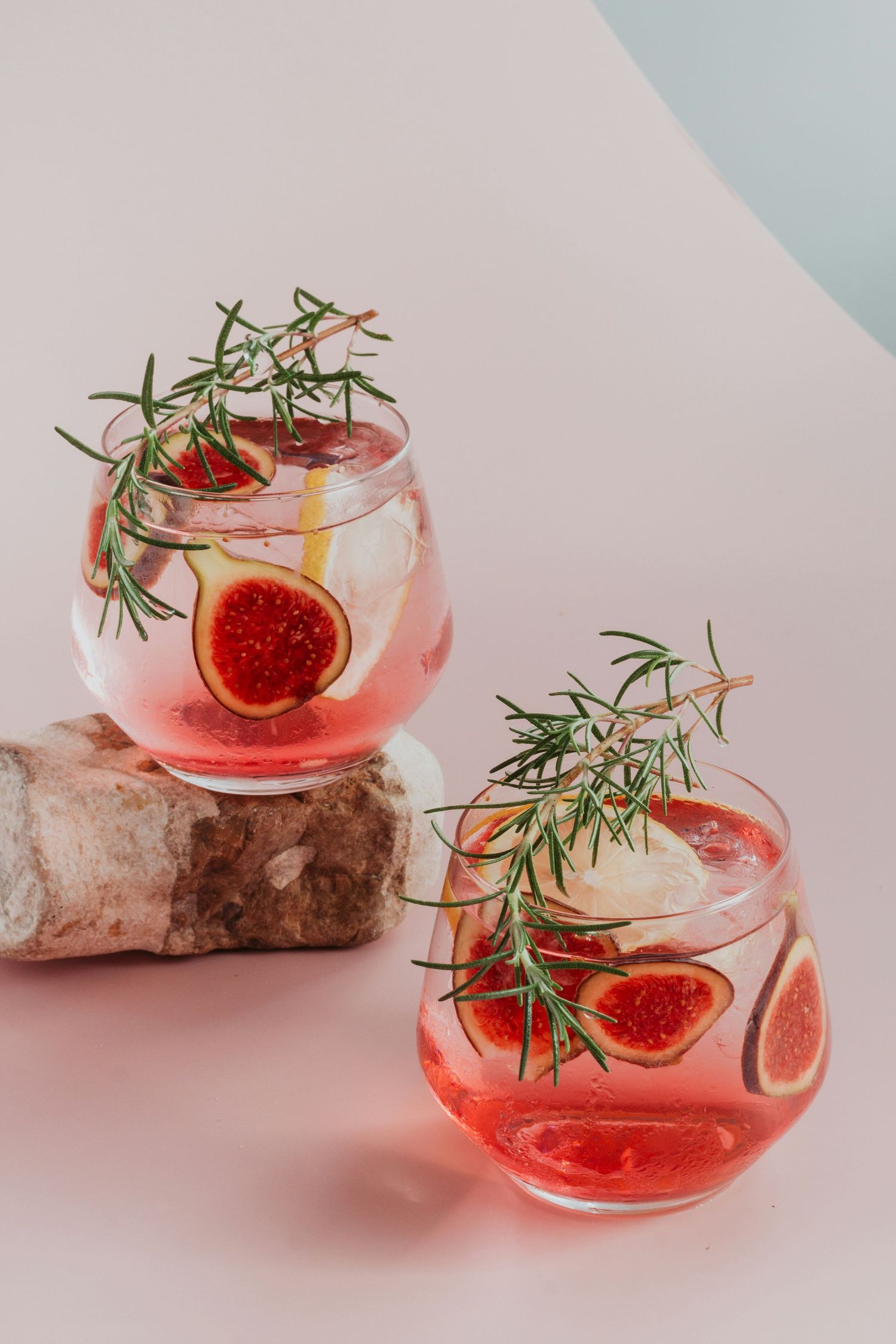 Two elegant stemless glasses with pink fig cocktails, garnished with fresh rosemary and ice, on a soft pink background.