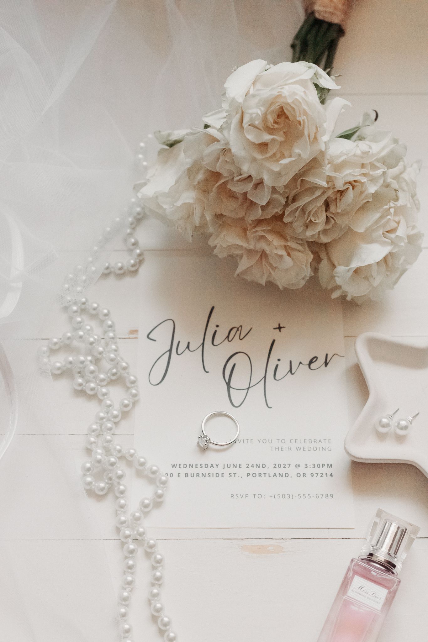 Elegant wedding flat lay with Julia and Oliver invitation, pearl necklace, ring, perfume, and white floral bouquet.