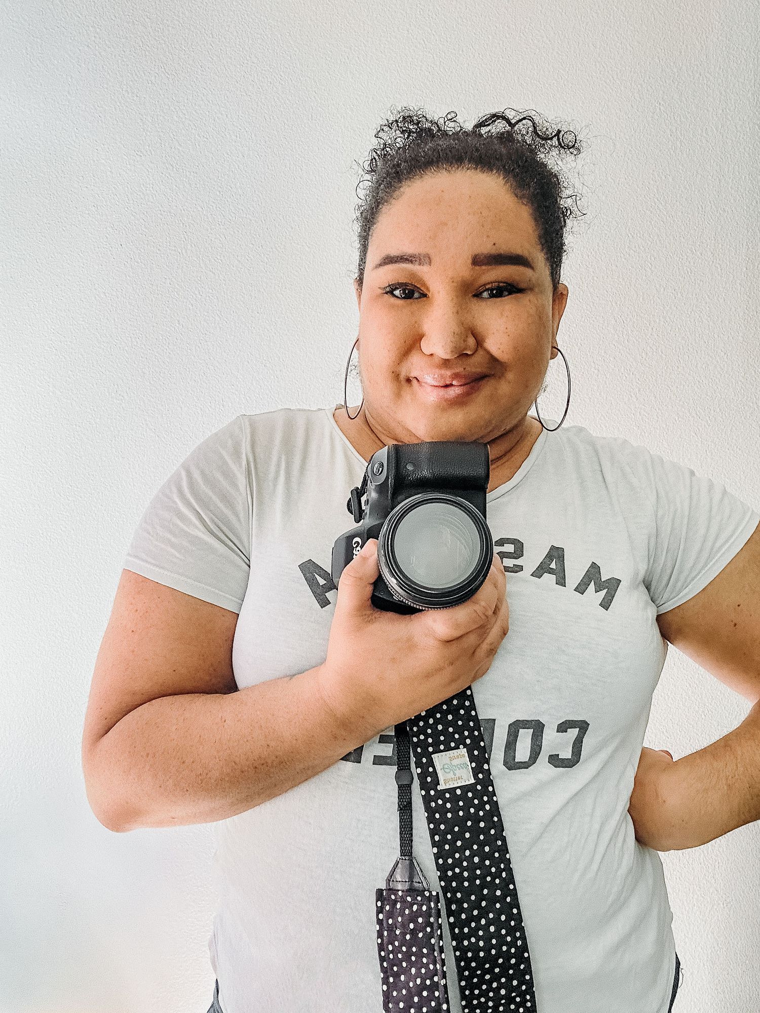 Woman in white graphic tee takes mirror selfie holding DSLR camera with polka dot strap, wearing hoop earrings.