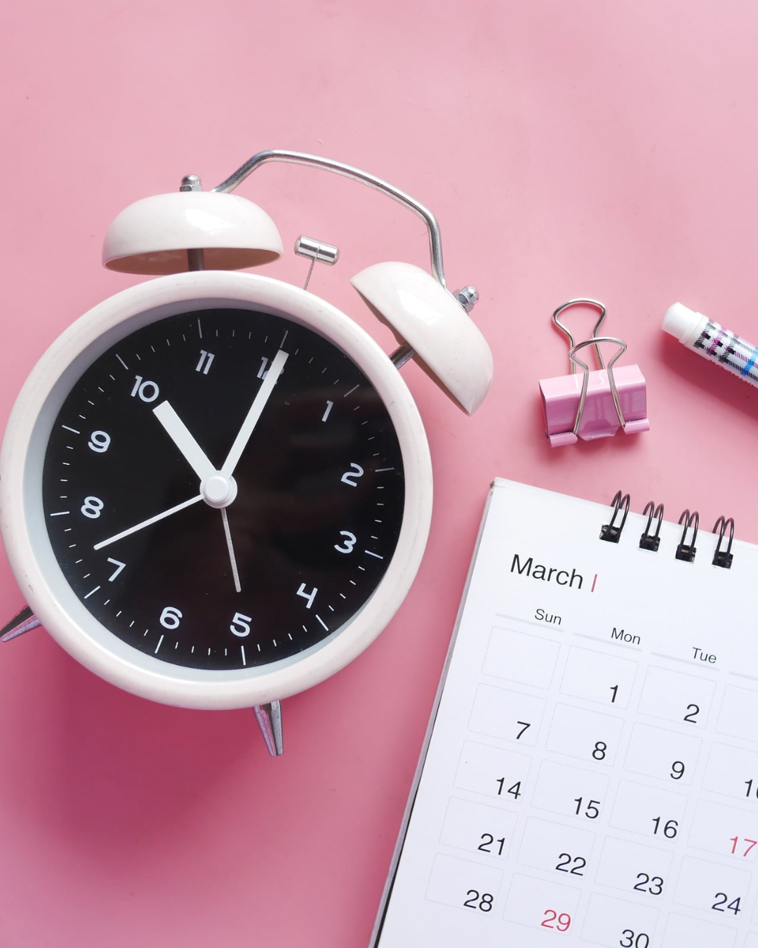 Pink alarm clock beside a March calendar, binder clips, and eraser on a pink background.