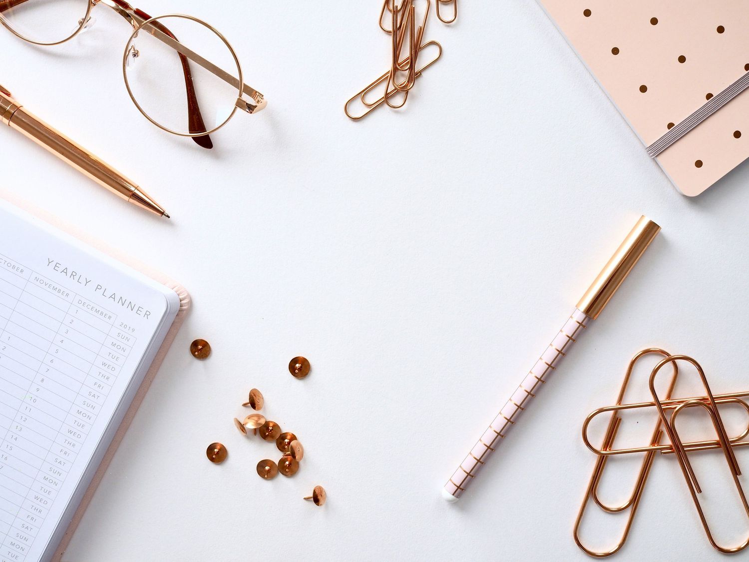Rose gold office supplies including glasses, pen, paper clips, and planner arranged on a clean white desk surface.