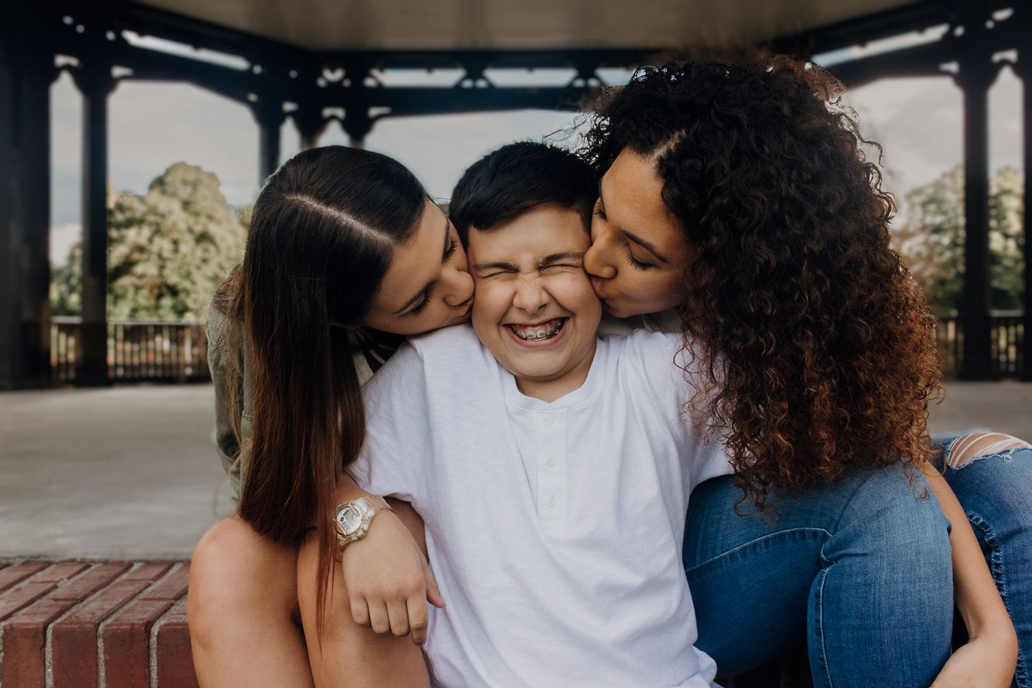 Two girls kissing a laughing boy on the cheeks while sitting together under a pavilion outdoors.