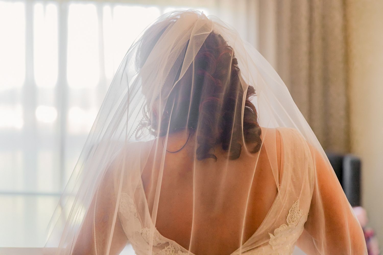 Bride in white wedding dress and veil seen from behind, with soft natural light from window.