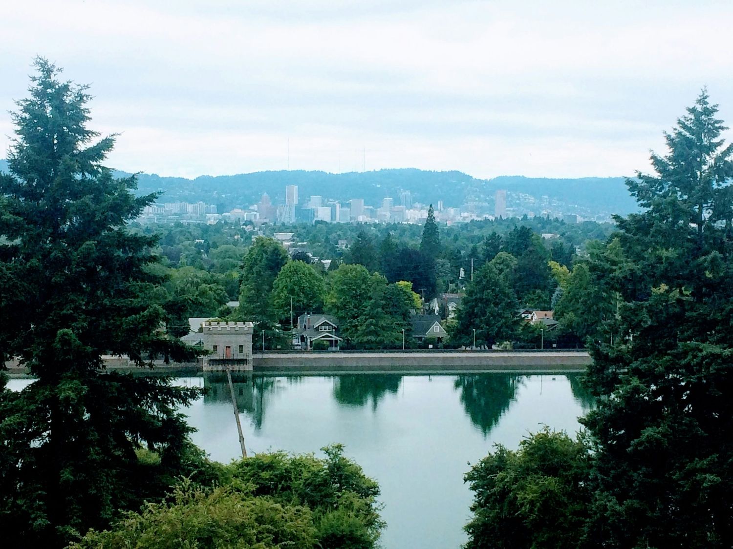 Scenic view of a city skyline beyond a calm lake surrounded by lush green trees and hills under an overcast sky.