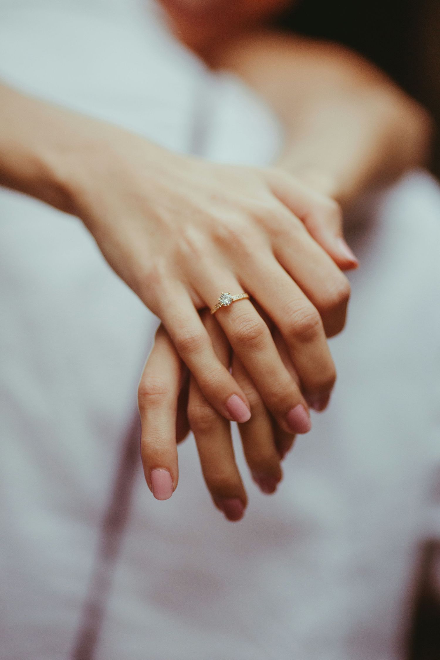 Close-up of female hands with a delicate gold diamond engagement ring on the ring finger, resting on white fabric.