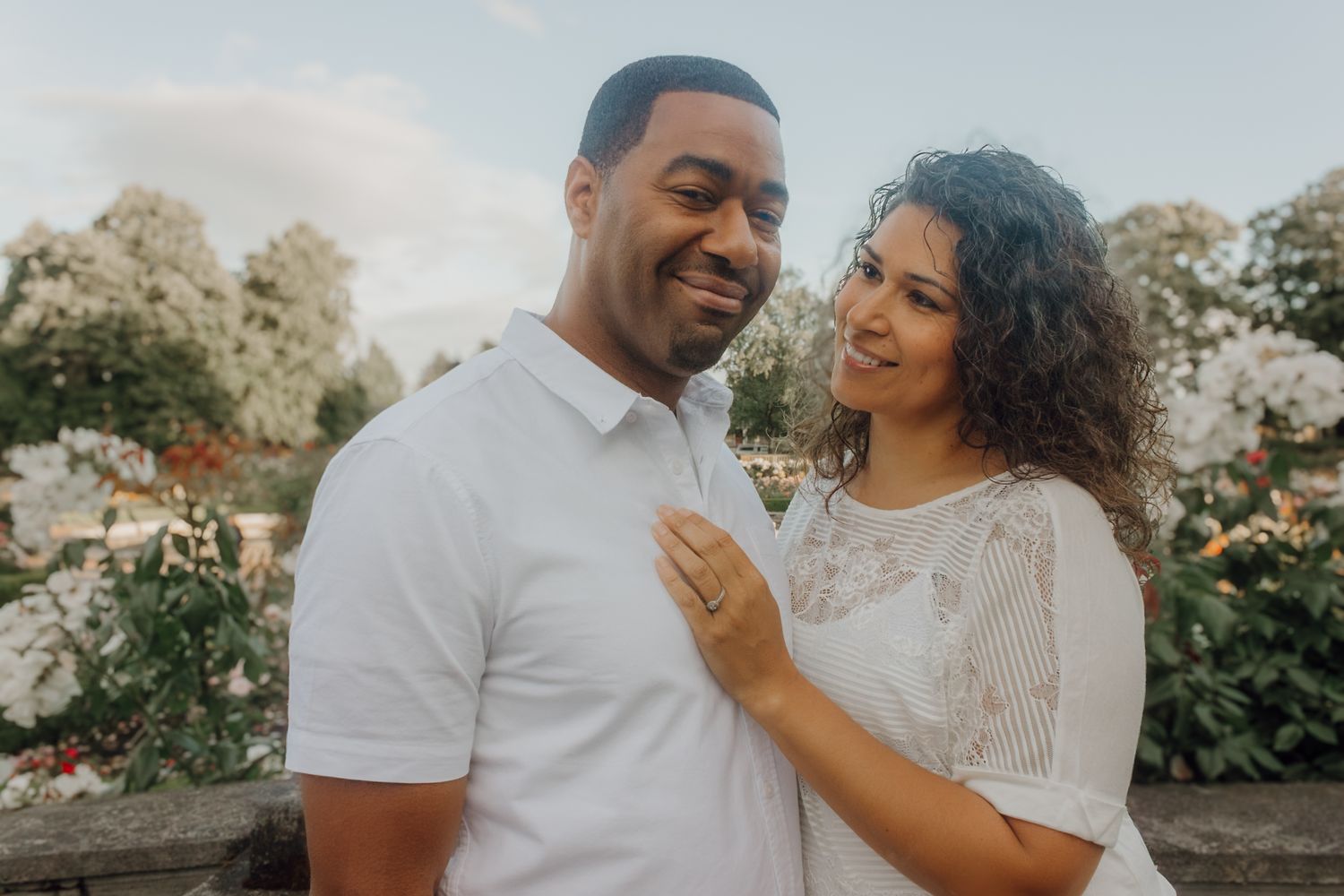 Couple in white outfits smiling at each other outdoors with lush green garden and flowers in background.