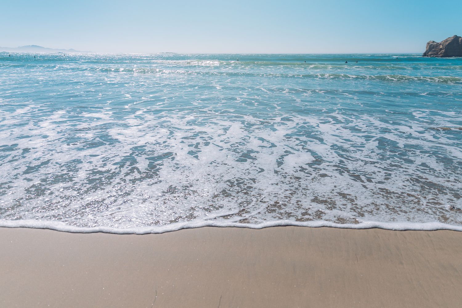 Gentle ocean waves washing onto a sandy beach with clear blue sky and turquoise water on a sunny day.