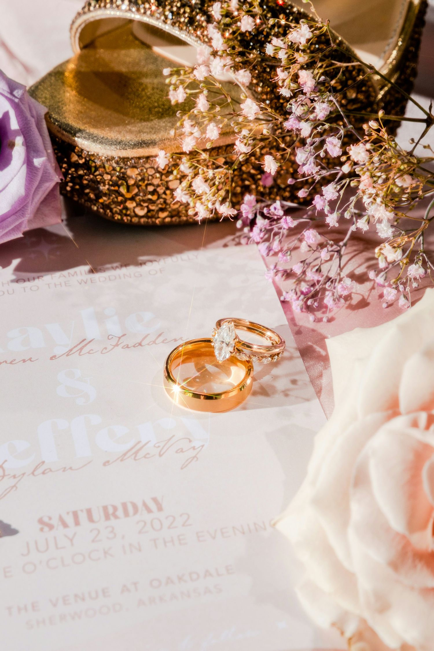 Gold wedding rings rest on a July 23, 2022 wedding invitation with pink flowers and gold heels at The Venue at Gaedales.