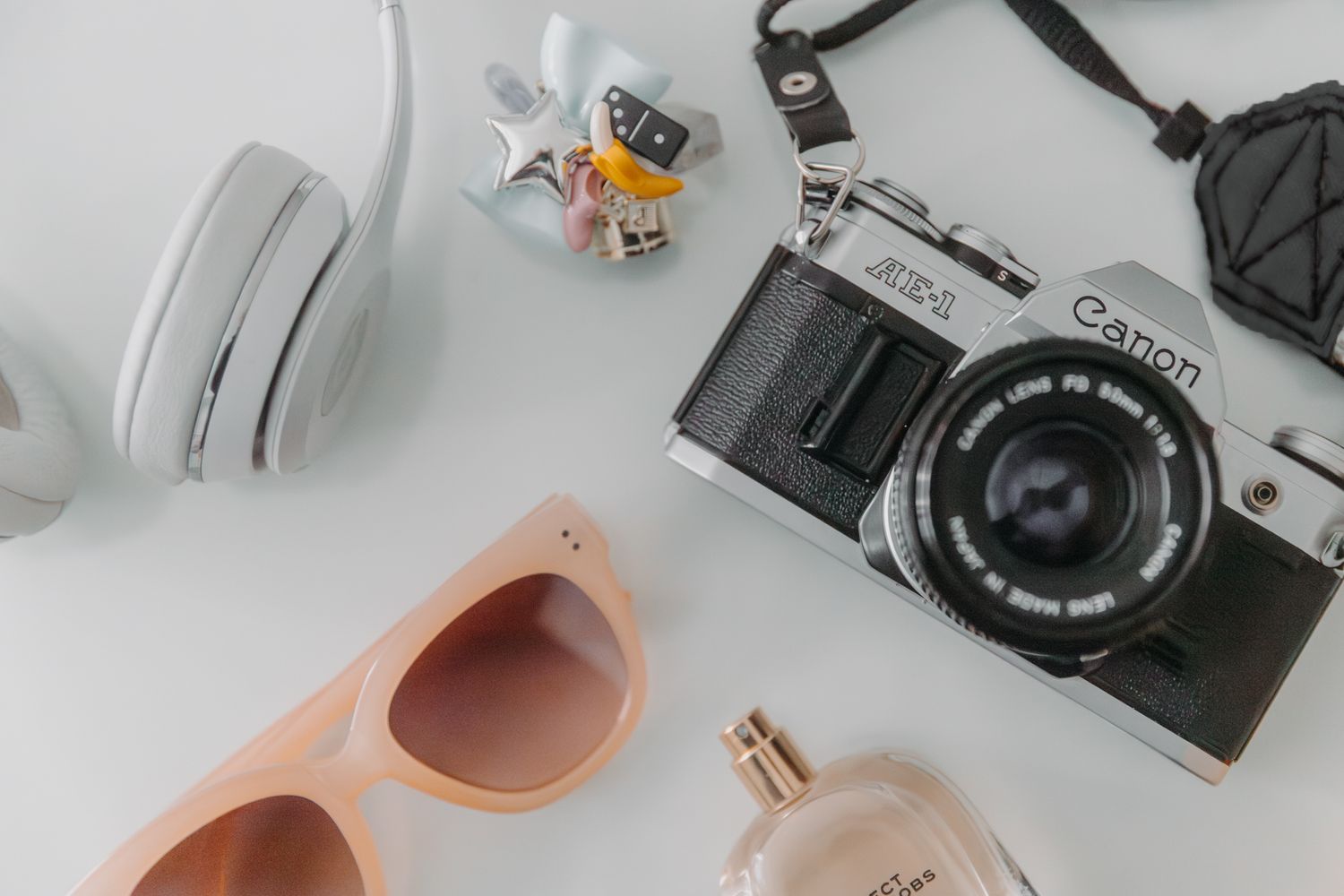 Flat lay of Canon film camera, white headphones, pink sunglasses, perfume bottle, and small toy on white surface.