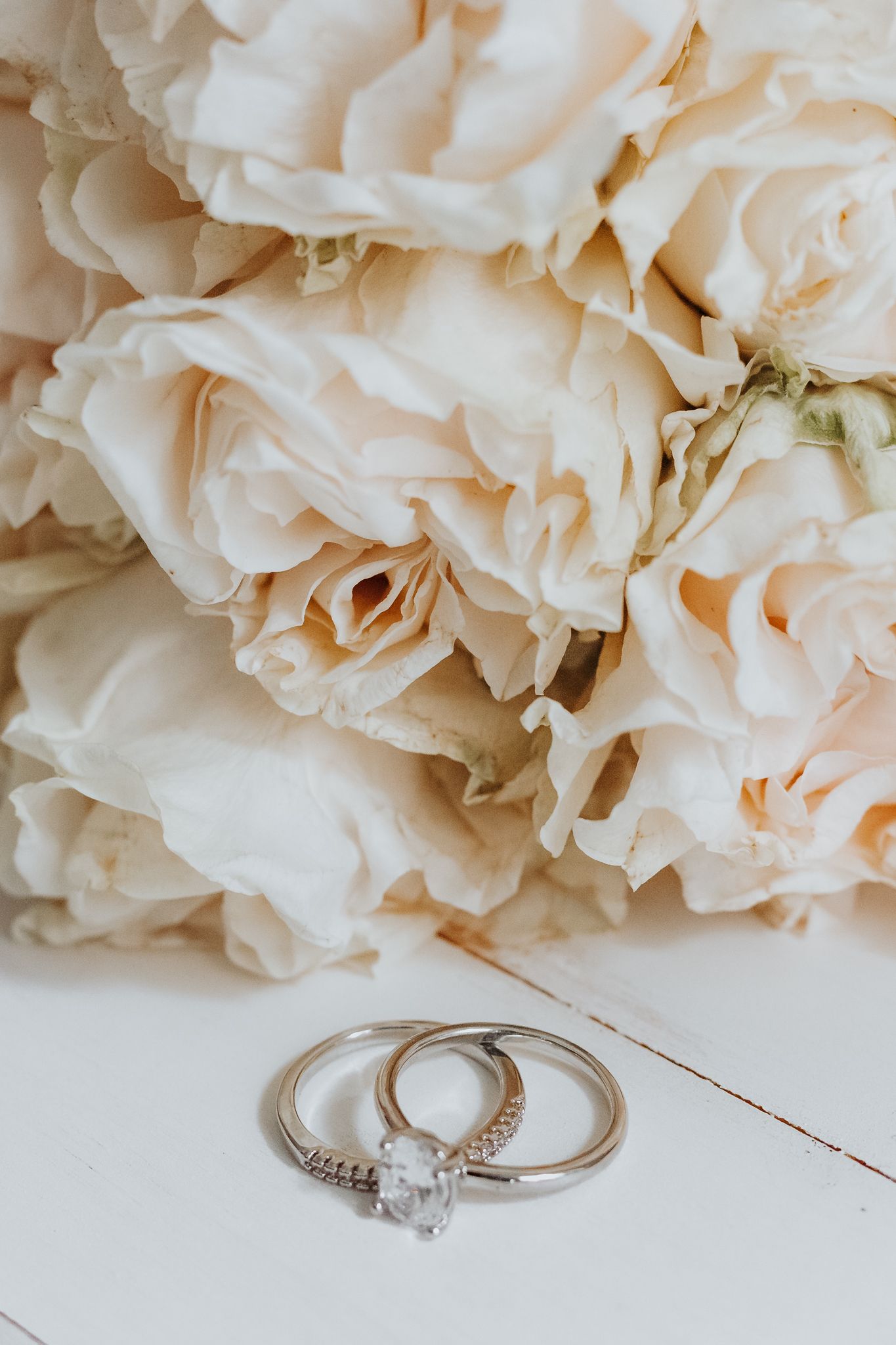 Silver wedding rings with diamond solitaire resting on white surface beside cream roses bouquet.