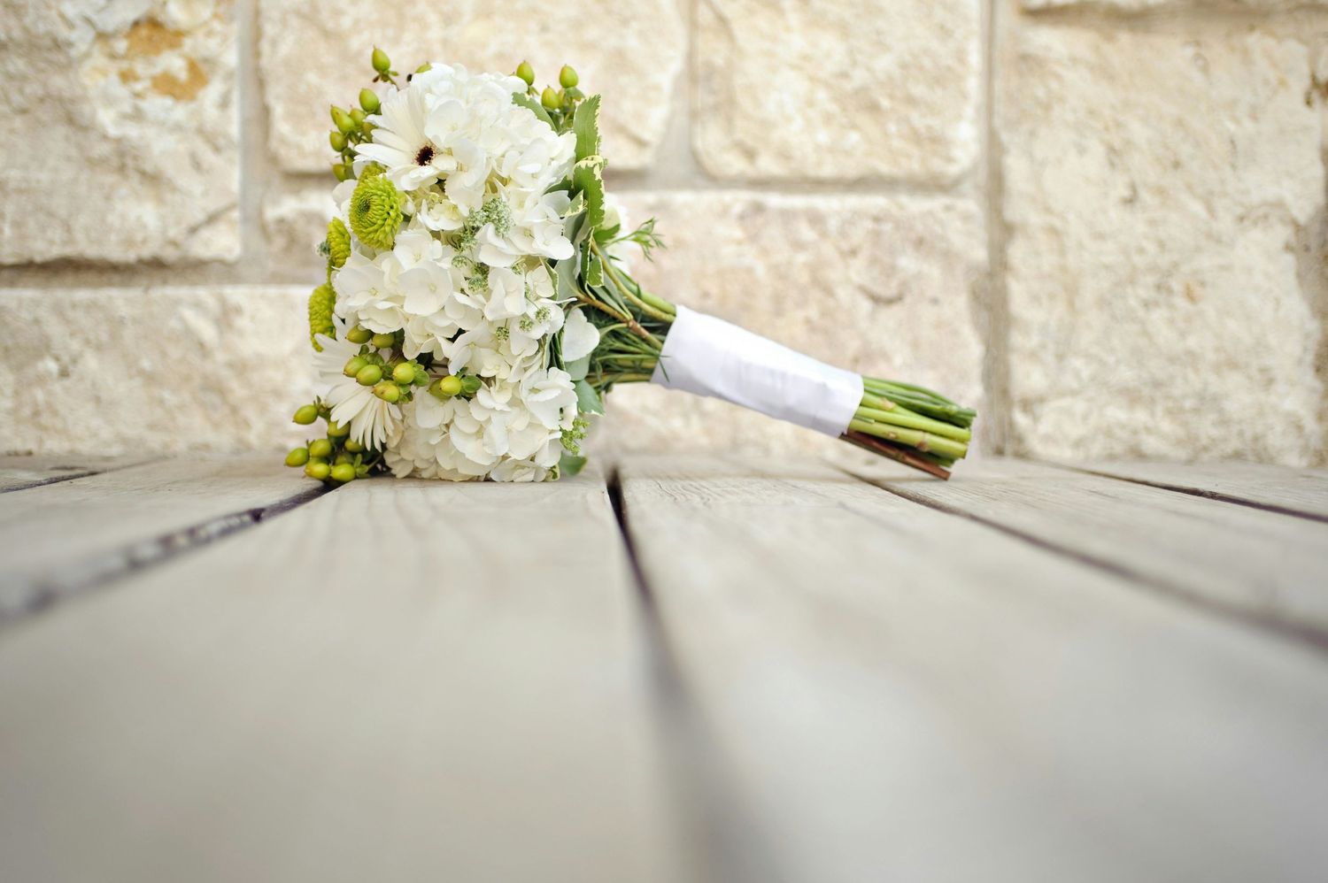 White wedding bouquet with green accents resting on wooden planks against a stone wall backdrop.