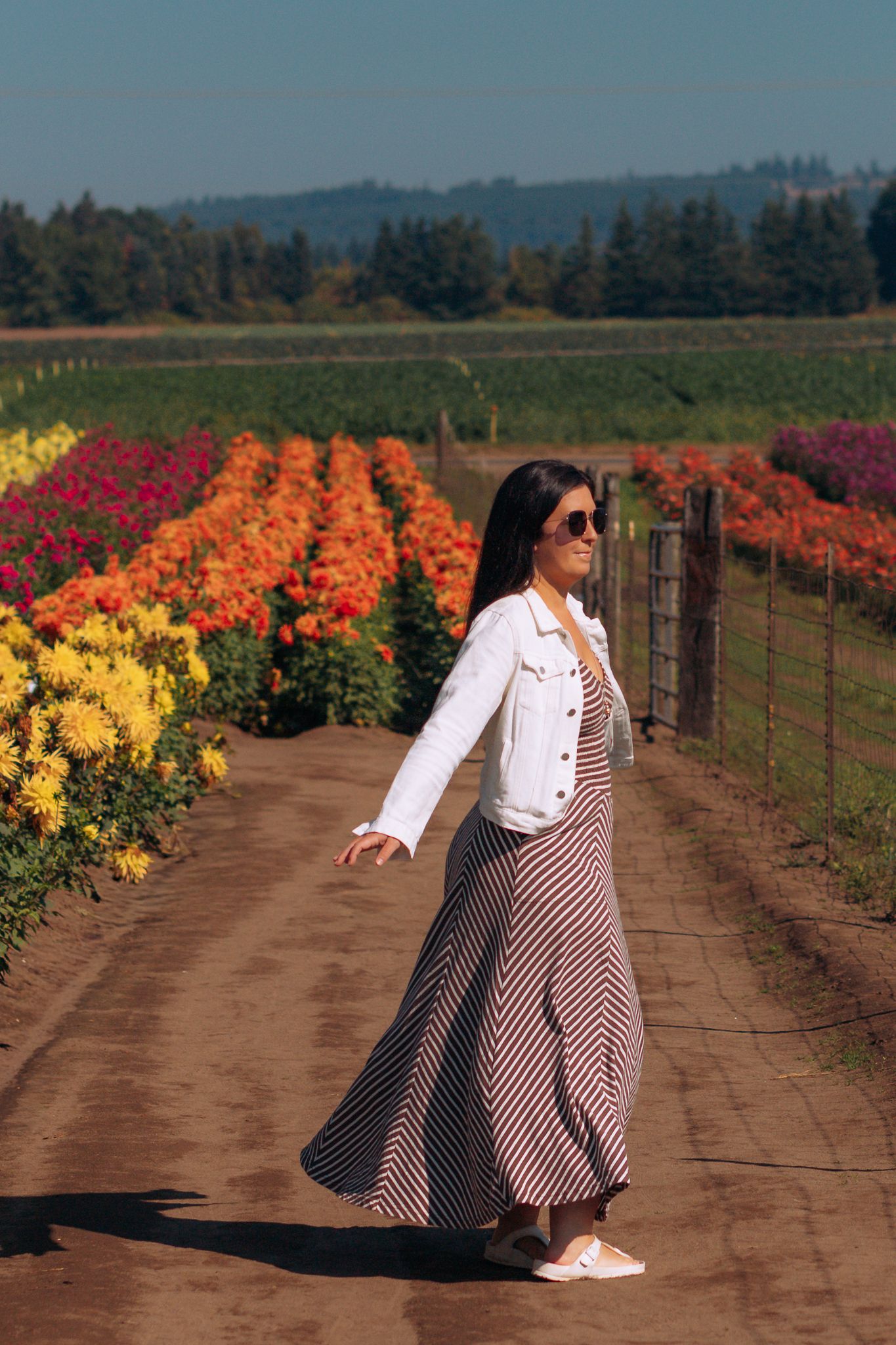 Woman in striped maxi skirt and white denim jacket walking along a dirt path beside colorful flower fields on a sunny day.