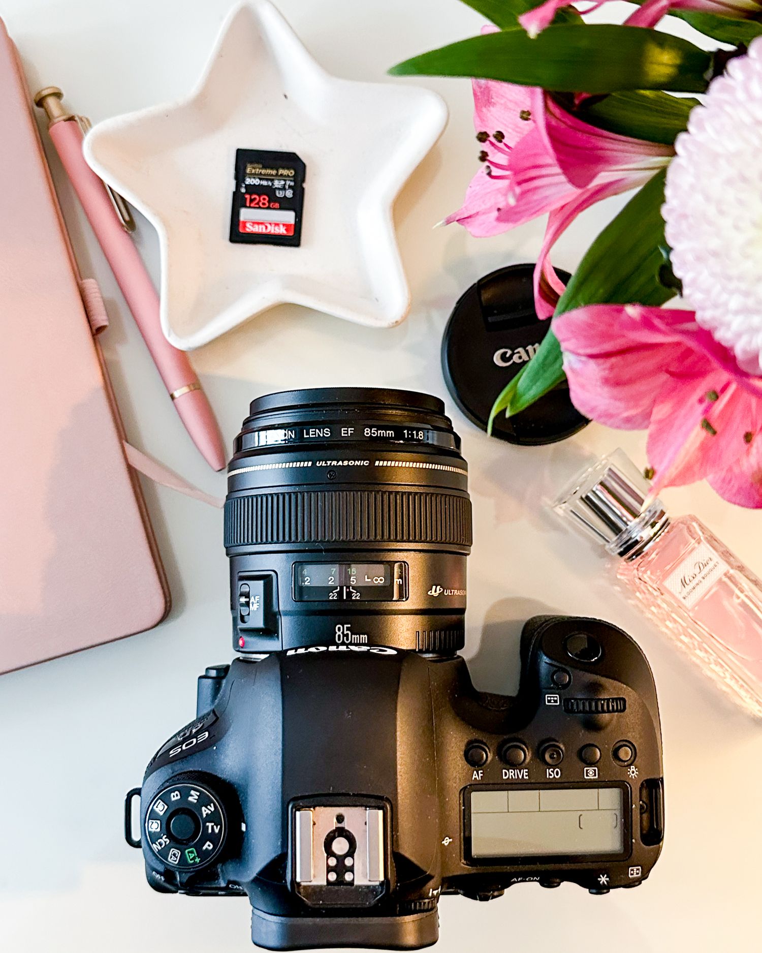 Canon DSLR camera with 85mm lens, SanDisk SD card, pink flowers, and Canon lens cap on white surface.