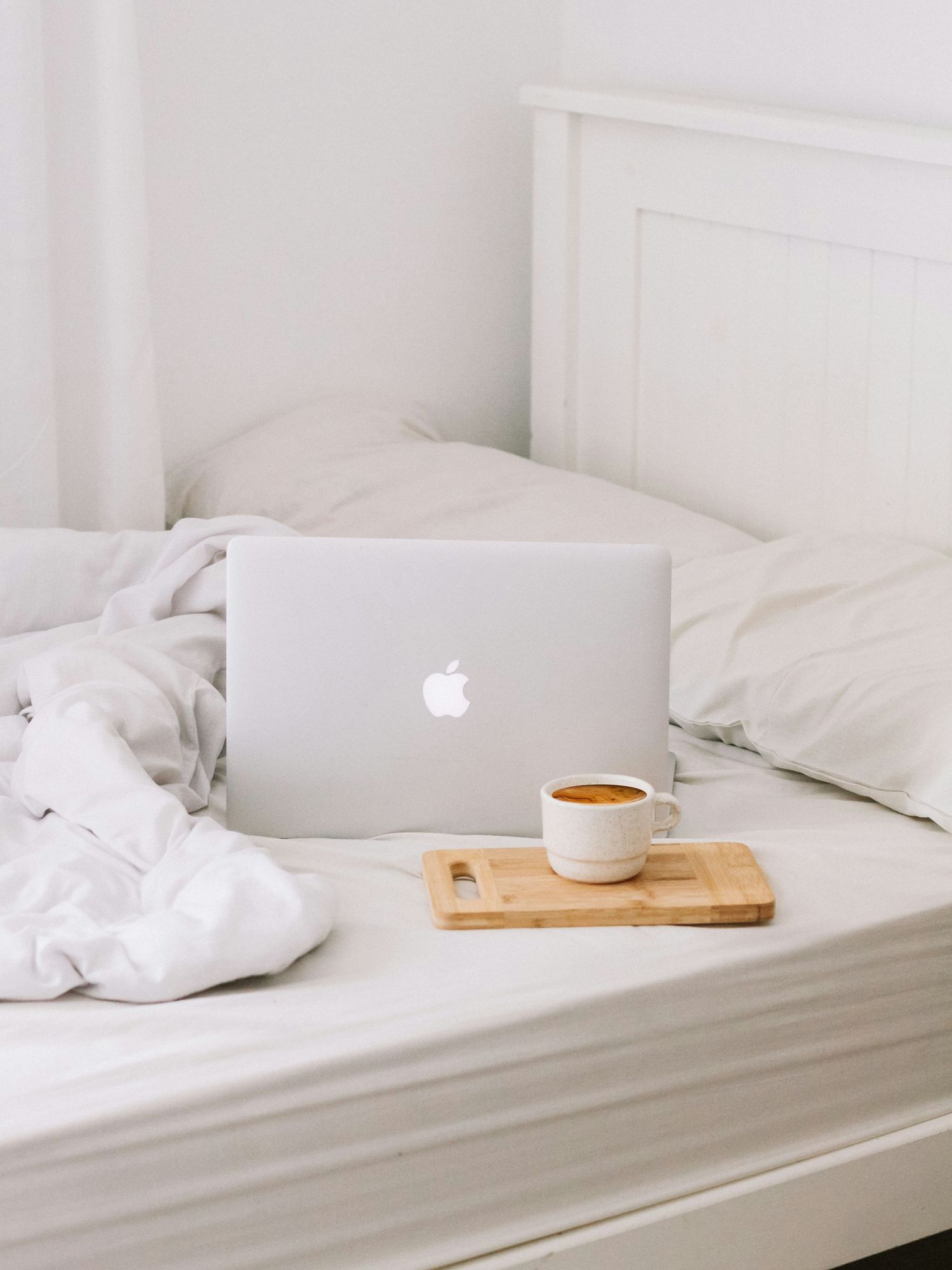 A MacBook laptop and a cup of coffee on a wooden tray rest on a neatly made white bed with white bedding.