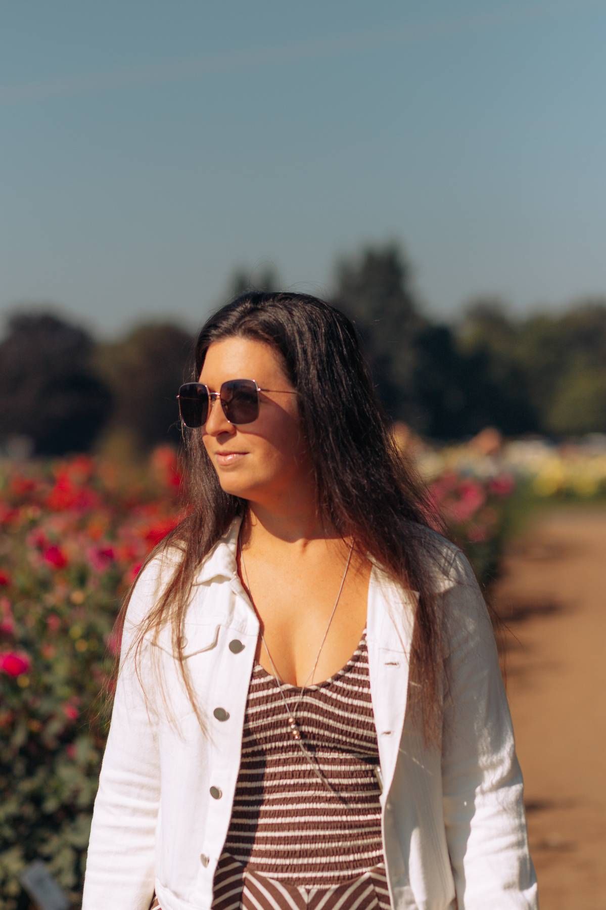 Young woman with long dark hair wearing sunglasses and a white jacket stands in a sunny flower garden.