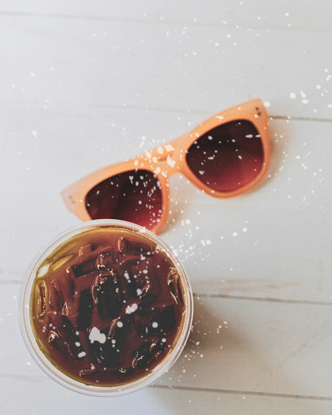 Sunglasses and a glass bowl filled with dark liquid on a white wooden surface with scattered salt or sugar crystals.