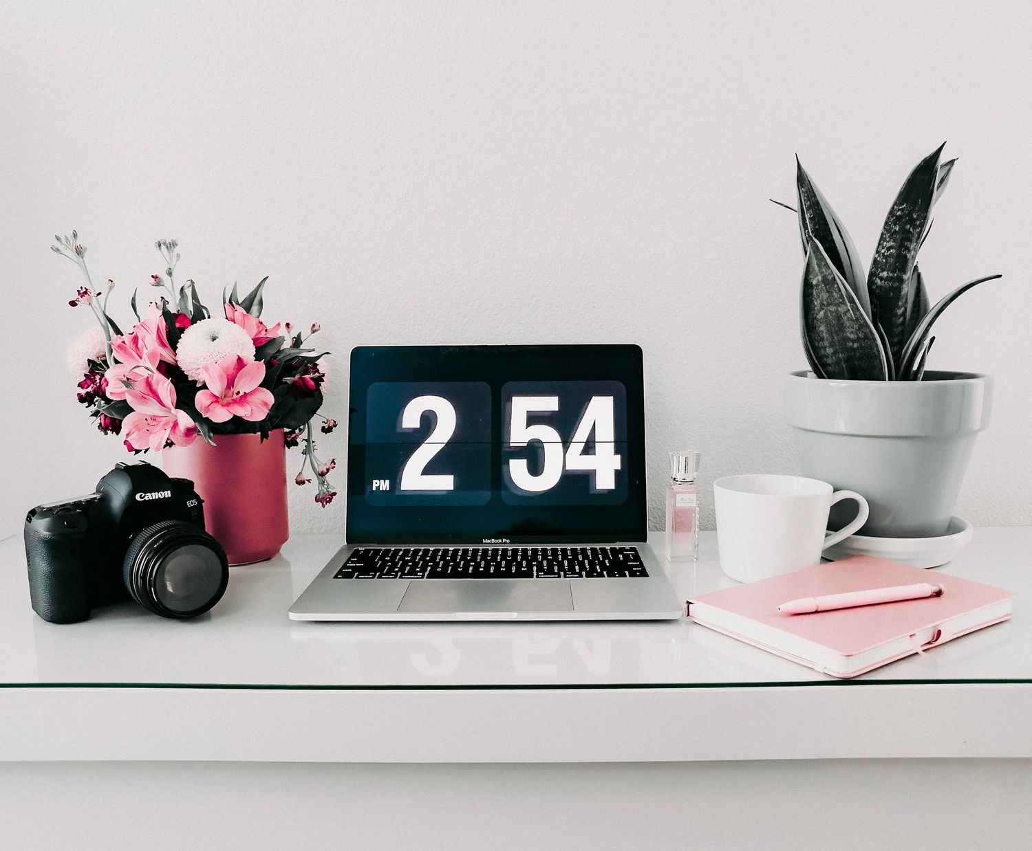 Minimalist desk workspace with pink and white decor, plants, laptop, and office accessories arranged on a white shelf.