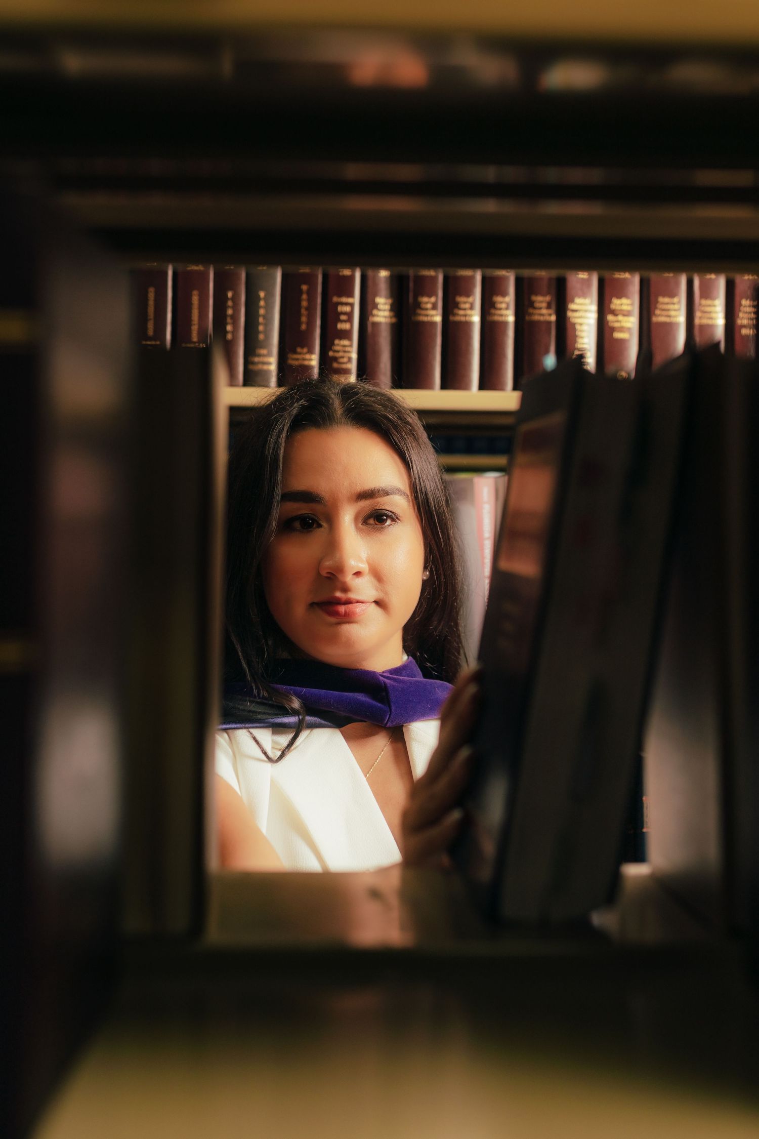 Graduate in white dress and purple stole seen through library bookshelf gap.
