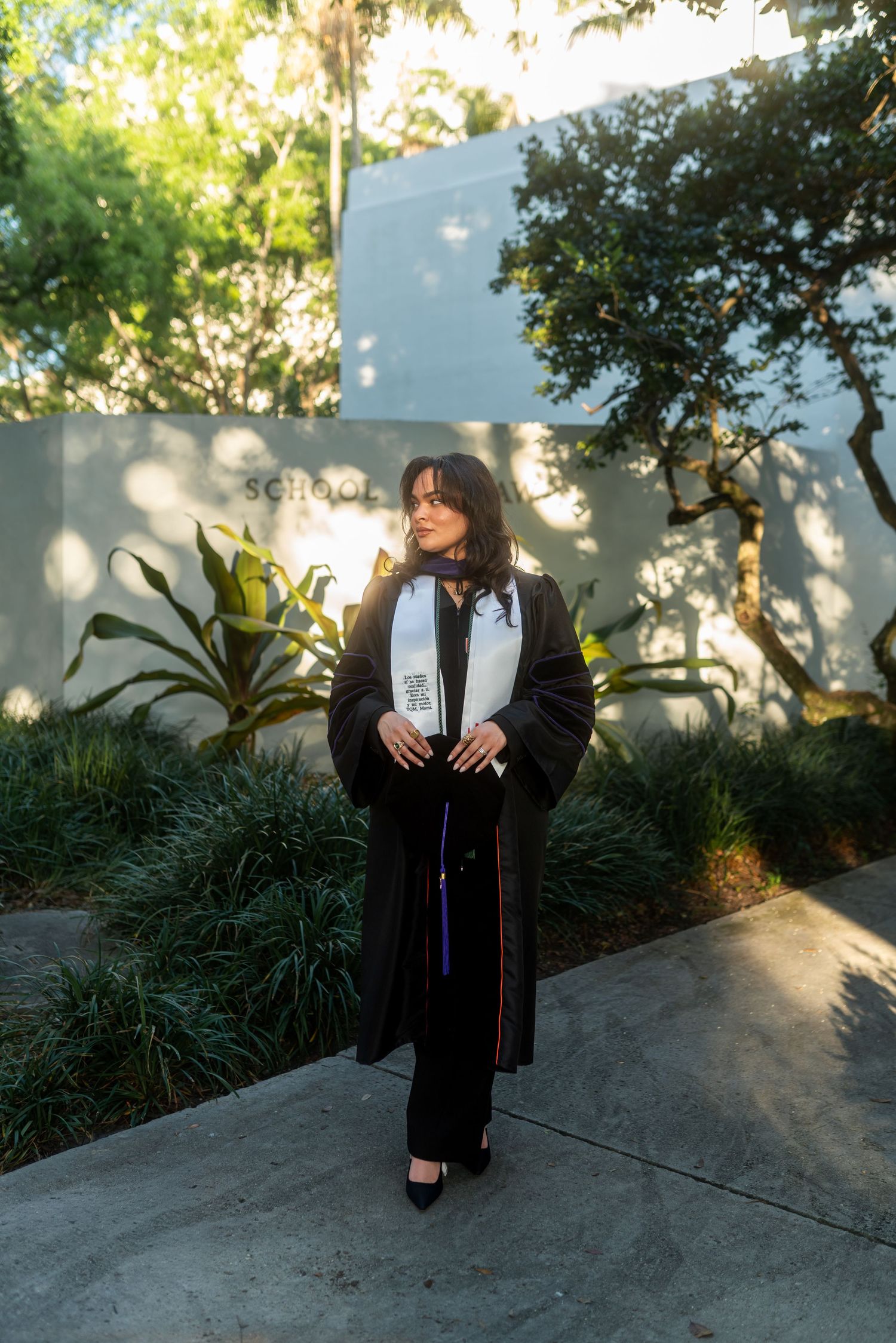 Graduate in black cap and gown poses outdoors near lush greenery and a school building on graduation day.