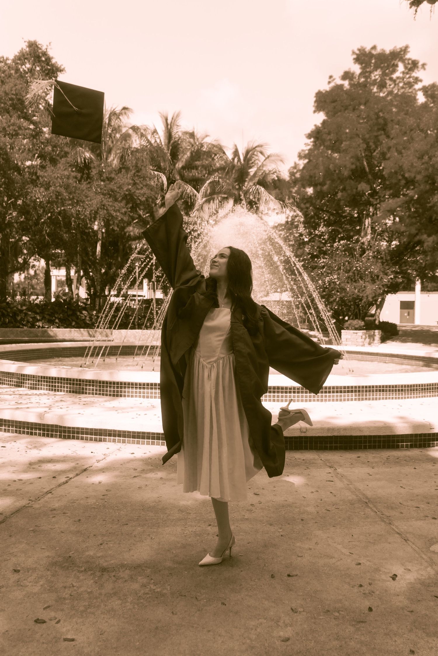 Graduate in black robe tosses cap near fountain in sepia-toned outdoor campus photo.